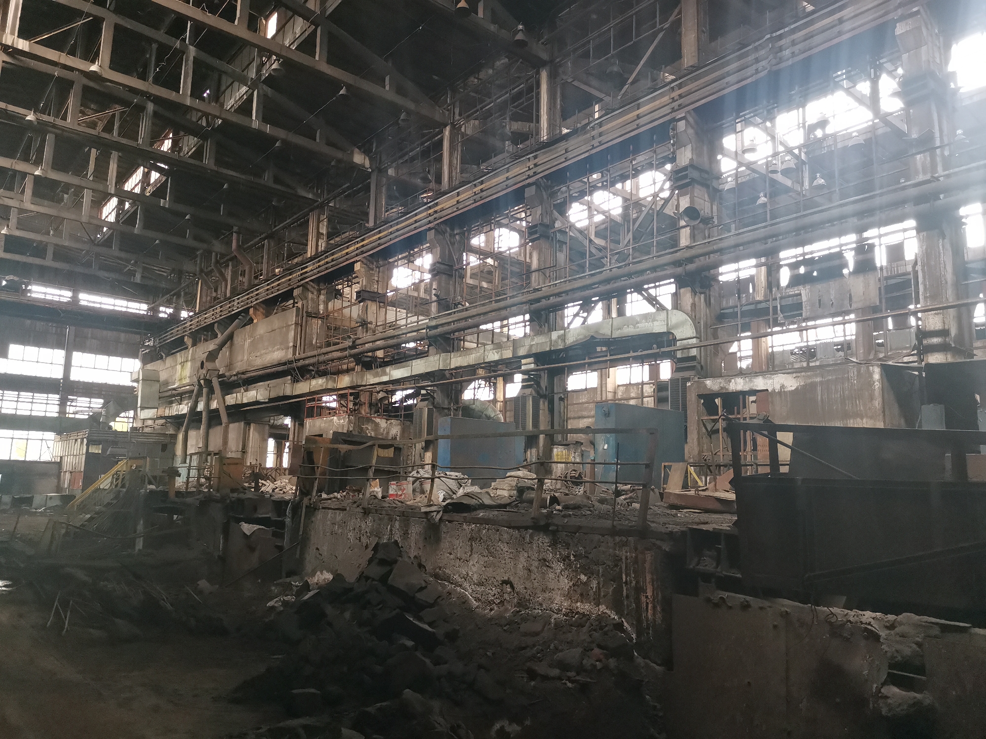 The image depicts the interior of a large, dilapidated industrial building, likely a former factory or plant, possibly located in Zhivkovo, Bulgaria. The structure is characterized by its high ceiling supported by a complex network of steel beams and girders. Natural light streams in through numerous windows, creating a stark contrast between the bright light and the dark, shadowy interior. The scene is cluttered with debris, scrap metal, and remnants of machinery, suggesting abandonment and decay. Various metal structures and piping run along the walls and ceiling, hinting at the building's past function. There are no people visible in the image. The overall impression is one of neglect and disuse.