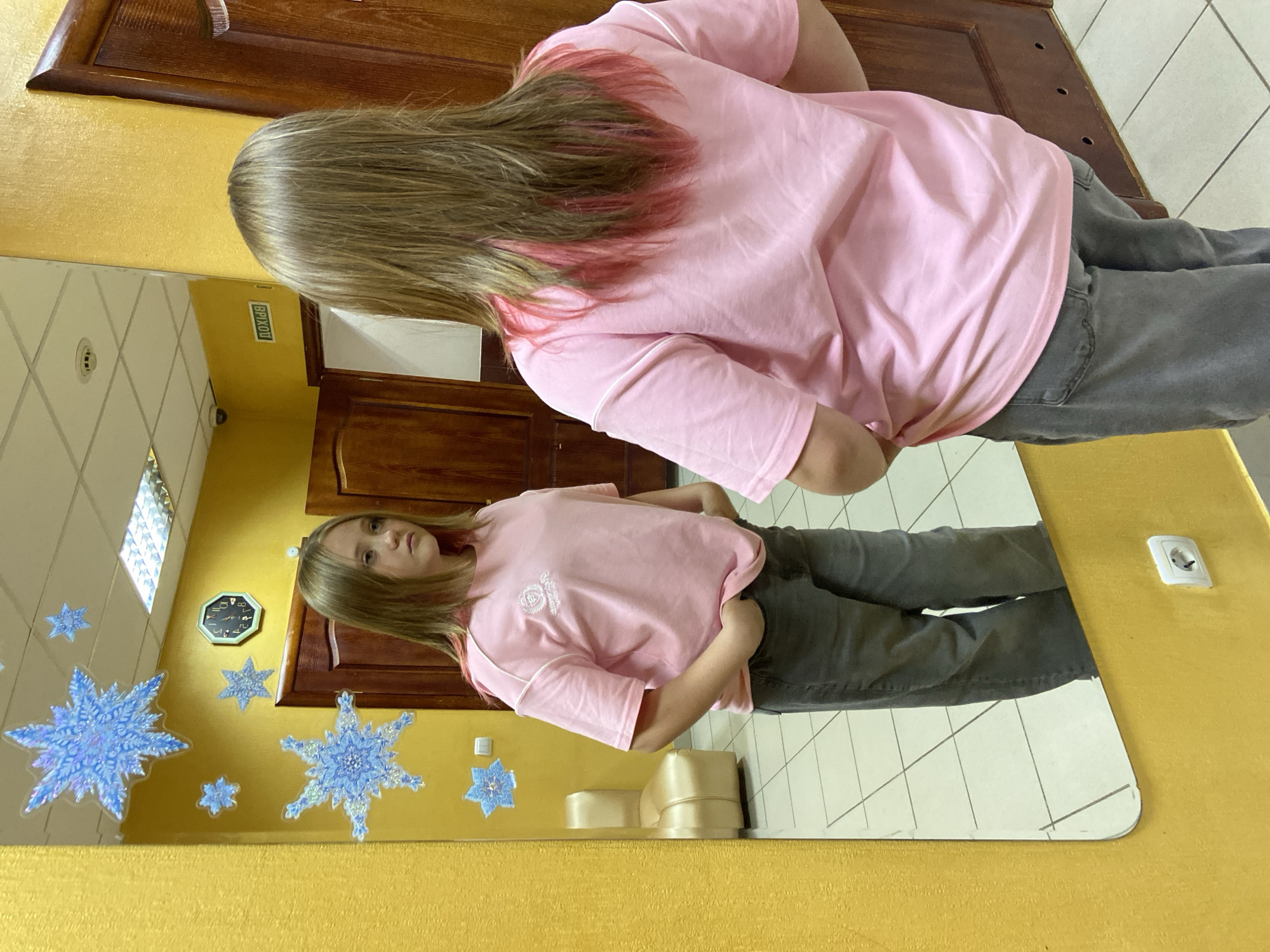 A young girl with long, blonde hair, partially dyed pink, stands in front of a large mirror. She is wearing a pink t-shirt with a crest on the left chest and dark gray pants. Her hands are tucked into her pockets. The mirror reflects the girl and the surrounding environment. The wall behind her is painted yellow and decorated with blue snowflake stickers. A round clock with Roman numerals is visible on the wall. To the right of the clock, a green exit sign with Russian text is illuminated. In the reflection, a cream-colored chair is visible. The floor is tiled with a grid pattern. The overall impression is that the photo was taken indoors, possibly in a public space or a home with decorative elements.