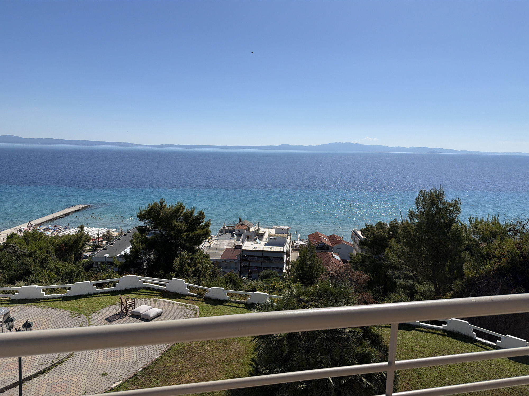 This is a wide shot of a coastal scene in Kassandra Municipal Unit, Greece, taken from a balcony or elevated vantage point. In the foreground, a white railing runs horizontally across the frame. Beyond the railing is a paved area with intricate brickwork, leading to a patch of grass. A simple wooden chair with two cushions is placed on the grass.
In the middle ground, lush green trees and vegetation create a natural barrier. Behind the trees, several buildings with white and red-tiled roofs are visible, suggesting a small town or resort area. Some of these buildings appear to be hotels or residential structures, with balconies and windows.
The background is dominated by the expansive, deep blue sea. The water is calm, with a gentle shimmer on its surface. In the distance, the hazy outline of a coastline with mountains is visible under a clear, bright blue sky. Closer to the shore, a long pier extends into the water, with several people visible on it and in the sea, indicating a beach area with swimmers and sunbathers. A section of beach with white umbrellas is also visible along the shoreline. A single bird is visible in the sky.