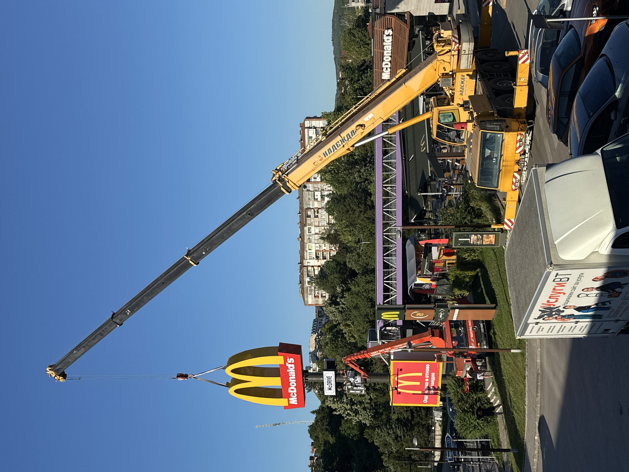 A large, yellow crane is in the process of lifting the iconic golden arches of a McDonald's sign, which is attached to a red base with the McDonald's logo and text. Below this, another, smaller orange crane is positioned, with its arm extended and supporting a portion of the main sign structure, possibly assisting in the installation or removal. Further down, a red truck with an extended arm is visible, also appearing to be involved in the operation. The McDonald's building is seen in the background, with its distinctive brown and yellow signage. Several cars are parked in a lot. In the foreground, a white truck with graphics on its side is parked. The scene is set outdoors, with a clear blue sky overhead and trees and apartment buildings in the distance, suggesting an urban environment. This event is taking place at the McDonald's restaurant located on Nikola Gabrovski Boulevard in Veliko Tarnovo, Bulgaria.