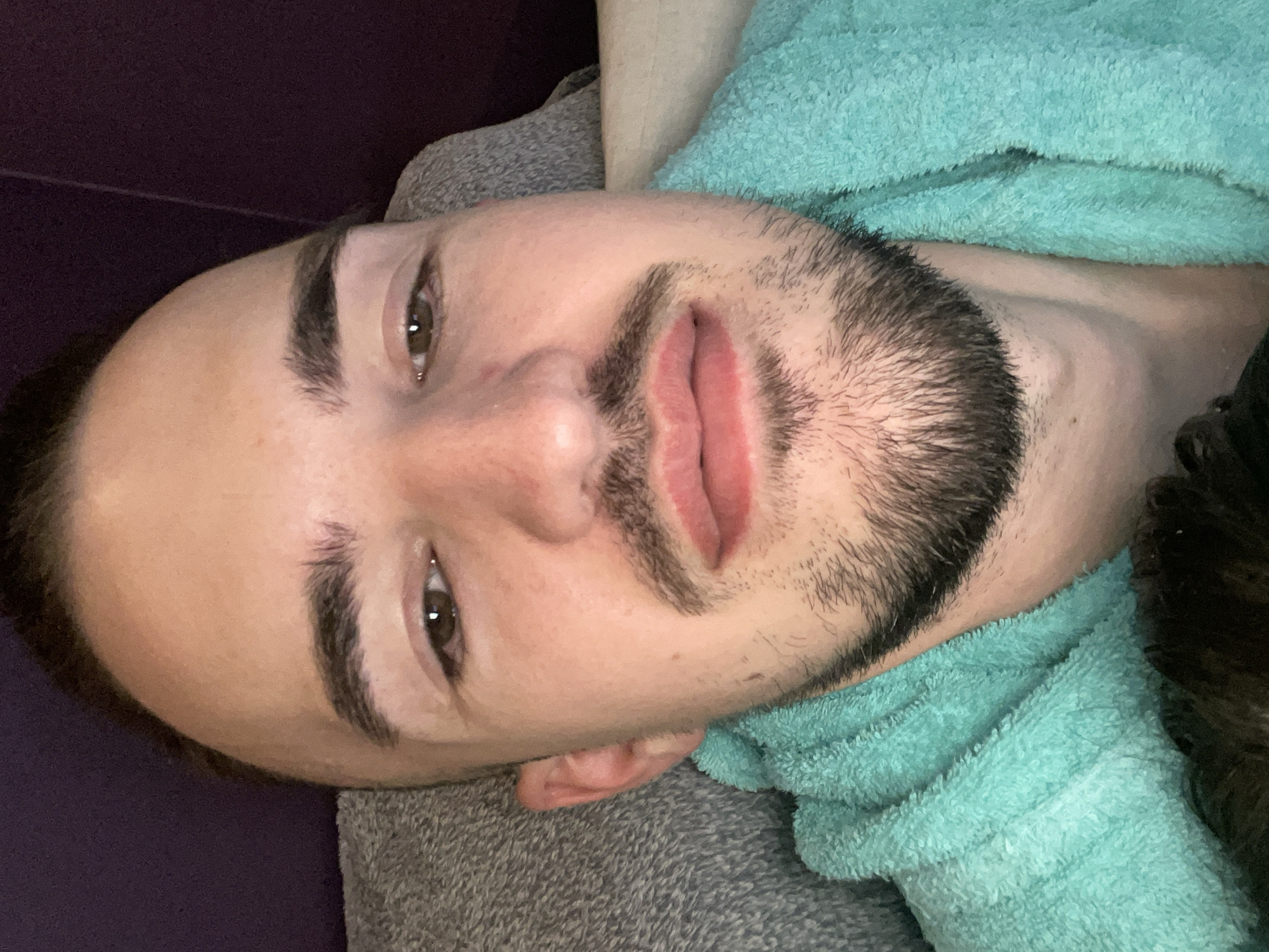 A close-up, horizontally oriented photo features a man's face. He appears to be lying down, with his head turned slightly to the left. His facial features are prominent: dark eyebrows, brown eyes, a nose, and full lips. He has a dark beard with stubble. The man seems to be resting on a gray fabric surface, possibly a pillow or blanket. He is wearing a light turquoise, possibly terry cloth, garment that covers part of his neck and shoulder. The background is a dark purple color in the top left corner, indicating a wall or similar surface. The image is indoors and the lighting suggests it could be in a room with controlled lighting. There is no indication or evidence of Sofia, Bulgaria in the image.