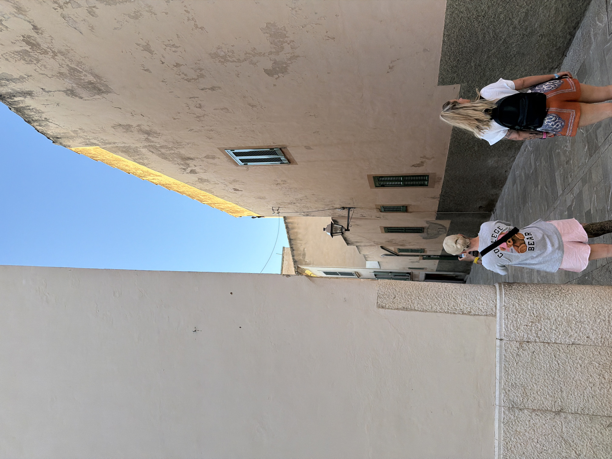 The image captures a narrow street in Alcúdia, Spain, framed by tall, weathered buildings. The left building has a smooth, light-colored facade, contrasting with the textured, beige wall of the building on the right, which shows signs of age and includes windows with closed shutters. Two women are walking along the street; the first, closer to the building with the windows, has blonde hair, a white shirt, patterned orange shorts, and a black backpack. The second woman, farther down the street, is wearing a white cap, a grey shirt with "COLLEGE BEAR" printed on the back, and pink shorts. The sky above the buildings is a clear blue.