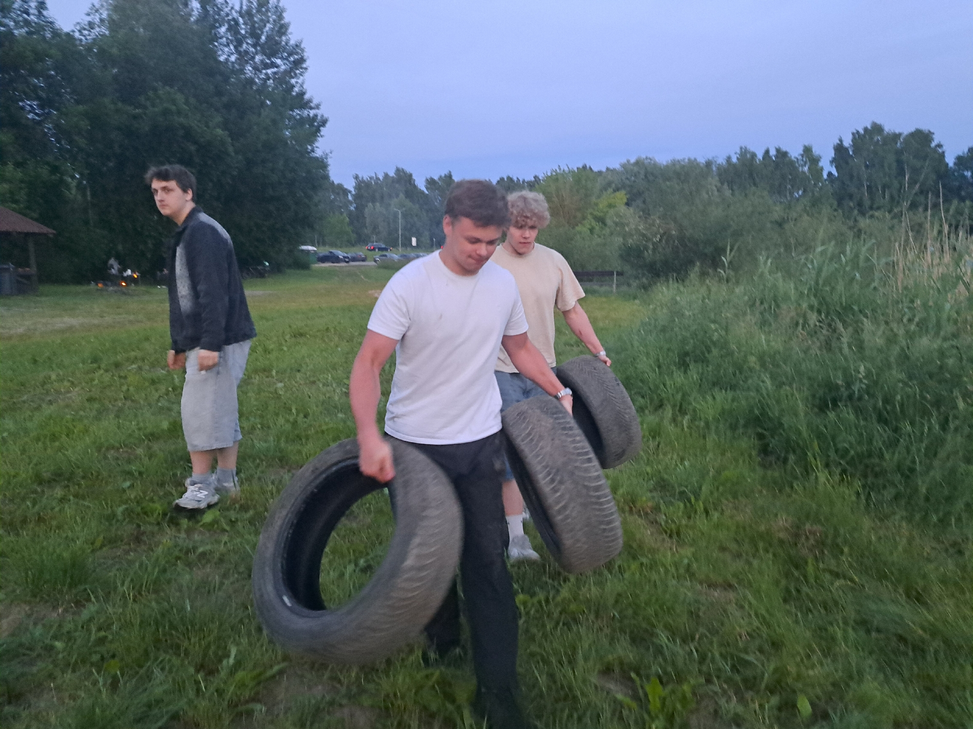 Three young men are in a grassy, outdoor area with trees in the background. The sky is overcast. In the foreground, two men are carrying car tires. The man on the left is wearing a white t-shirt and black pants, and he is carrying one tire. The man behind him is wearing a tan t-shirt and is carrying a second tire. To the left of the frame, another man is standing and looking towards the camera. He is wearing a dark jacket over a grey shirt and light-colored shorts. In the background, there are cars and a small wooden structure, suggesting a park or recreational area. The overall setting appears to be a rural or semi-rural location.