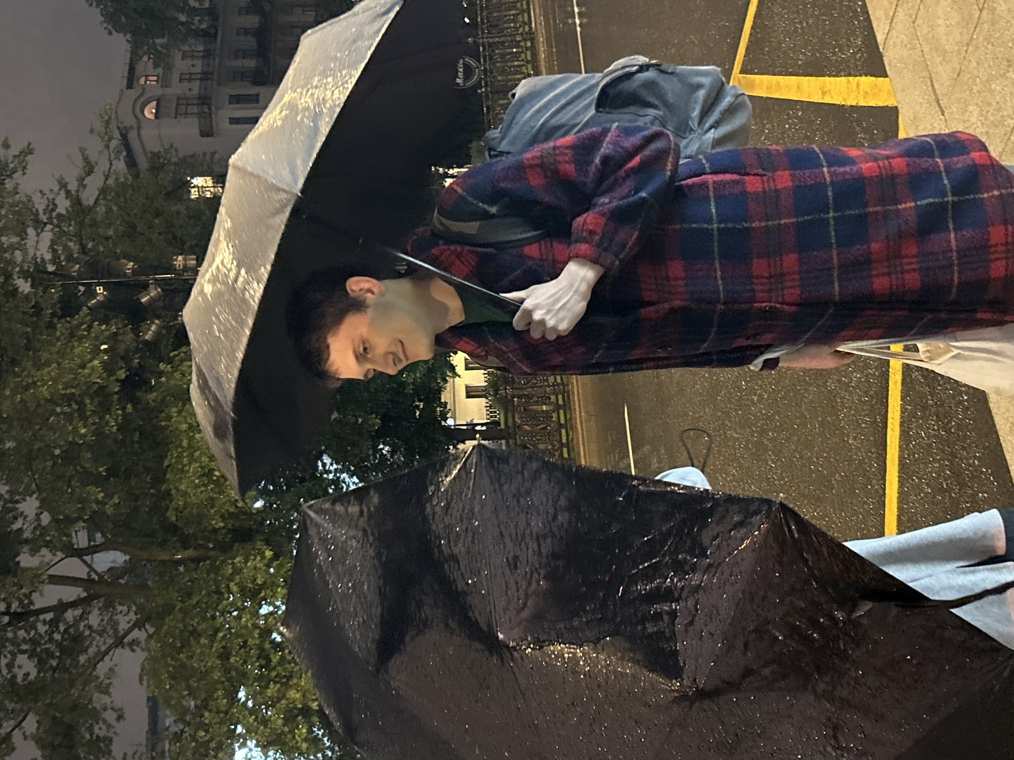 In the city of Unknown, Unknown, during a rainy evening, a young man with short brown hair, wearing a red and blue plaid coat over a green shirt, stands under a large black umbrella. He is holding the umbrella's handle with his right hand and a white tote bag in his left. A grey backpack is visible over his shoulders. To his left, partially obscuring him, is another large black umbrella, appearing to shelter a person wearing a light blue garment. The ground is wet, reflecting the ambient light from street lamps and nearby buildings, indicating it has recently rained or is currently raining. A yellow line is painted on the wet asphalt surface, suggesting a pedestrian crossing or a road marking. In the background, trees with green foliage are present, and a multi-story building with arched windows and balconies is visible. The overall scene depicts a common urban moment of people navigating a rainy night.