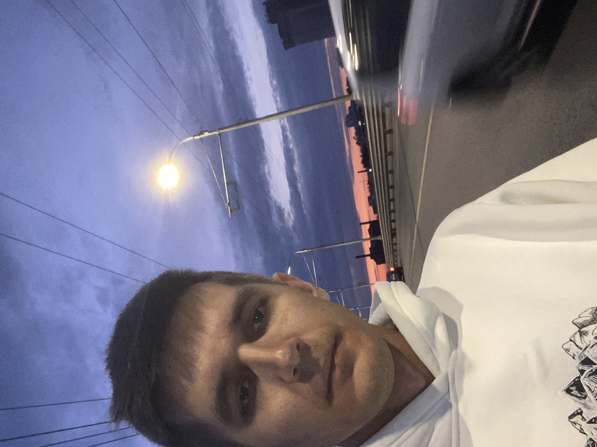 A young man in a white hoodie stands on a bridge in Novokuybyshevsk, Russia, as dusk settles. The sky is a deep purple, transitioning to orange at the horizon, with dark clouds visible. A streetlamp illuminates the scene, casting a warm glow. Power lines crisscross the sky. In the background, the silhouette of a city skyline, including industrial structures and buildings, is visible. A car, blurred by motion, passes on the road. The man is looking directly at the camera, his expression neutral. The bridge railing runs along the edge of the road.
