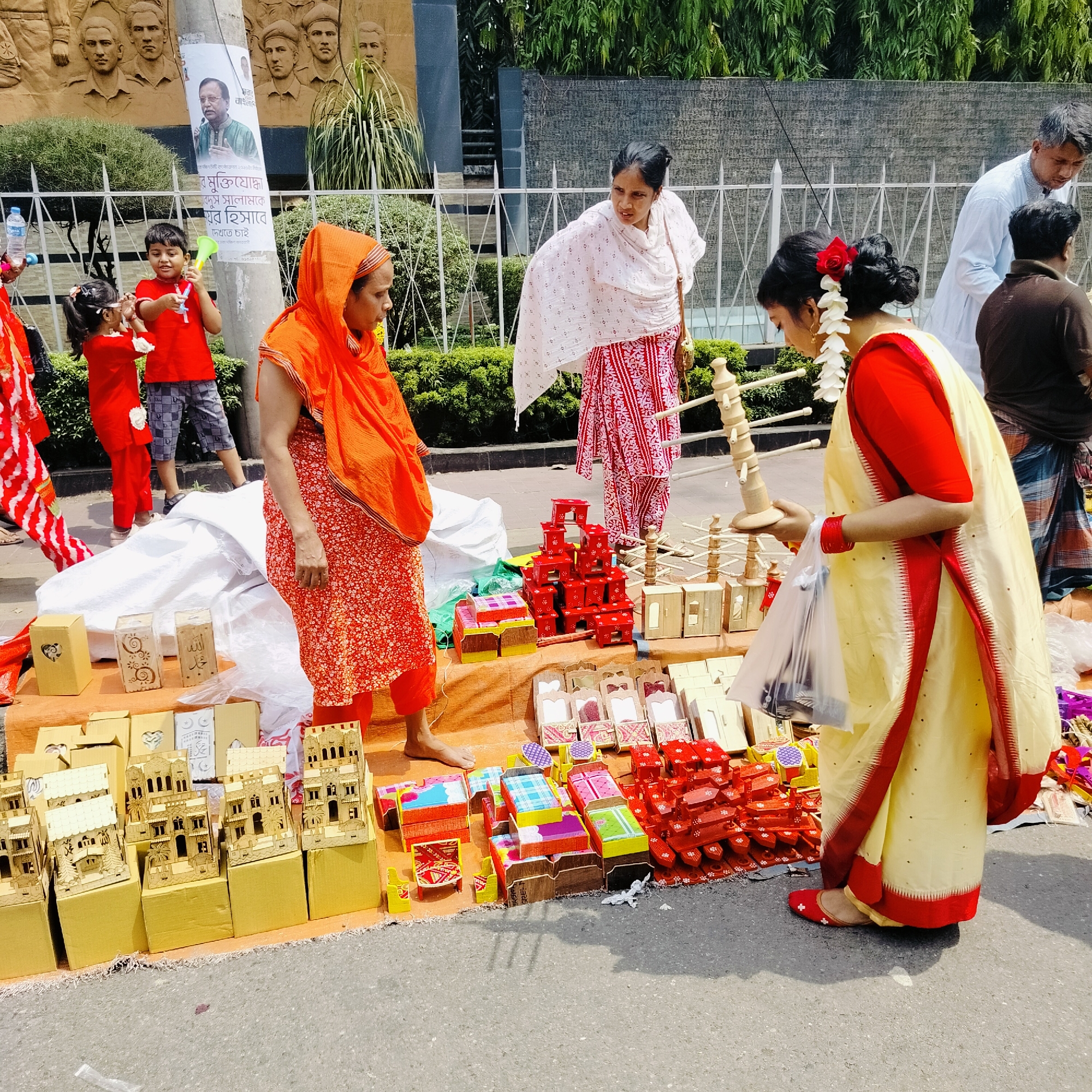 A daytime street scene in Dhaka, Bangladesh, shows two women examining items for sale from a street vendor. The vendor, standing to the left, is an older woman wearing a red and white patterned sari with an orange shawl. A woman in a cream-colored sari with red trim stands to the right, holding a wooden stand. A third woman, wearing a red and white patterned shalwar kameez with a white shawl, stands behind the vendor. Two children, a boy and a girl, stand in the background; both are wearing red shirts, with the boy in shorts and the girl in pants. Various wooden items, including miniature houses and furniture, are displayed on a mat. Behind the group is a memorial wall with bas-relief figures and a sign in Bengali, partially obscured by a utility pole. A man in a white shirt is visible on the right. The setting appears to be a public area near a monument or memorial site, as suggested by the architectural elements in the background.