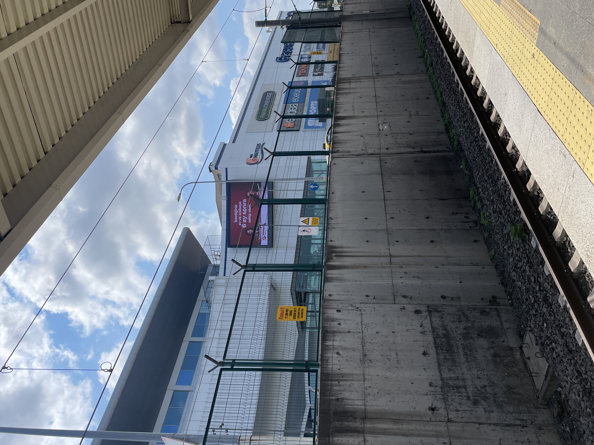 The image depicts a view from a train station platform in Kadıköy, Turkey. In the foreground, a yellow tactile paving strip runs along the edge of the platform, bordering a railway track. A concrete wall, showing signs of wear and tear, forms the boundary between the platform area and the building behind it. Above the wall, a green metal fence encloses a wide, white-colored building. This building features multiple windows and several large signs displaying various brand names such as "NAUTILUS," "PARIBUCINEVERSE," "endea," "ebebe," "SPORTS," "TEKNOSA," and "Gratis." A large digital billboard on the left side of the building displays an advertisement with text in Turkish and an image of a person. The sky above is partly cloudy with a mix of blue sky and white clouds. Overhead, power lines are visible, suspended by poles and attachments.
