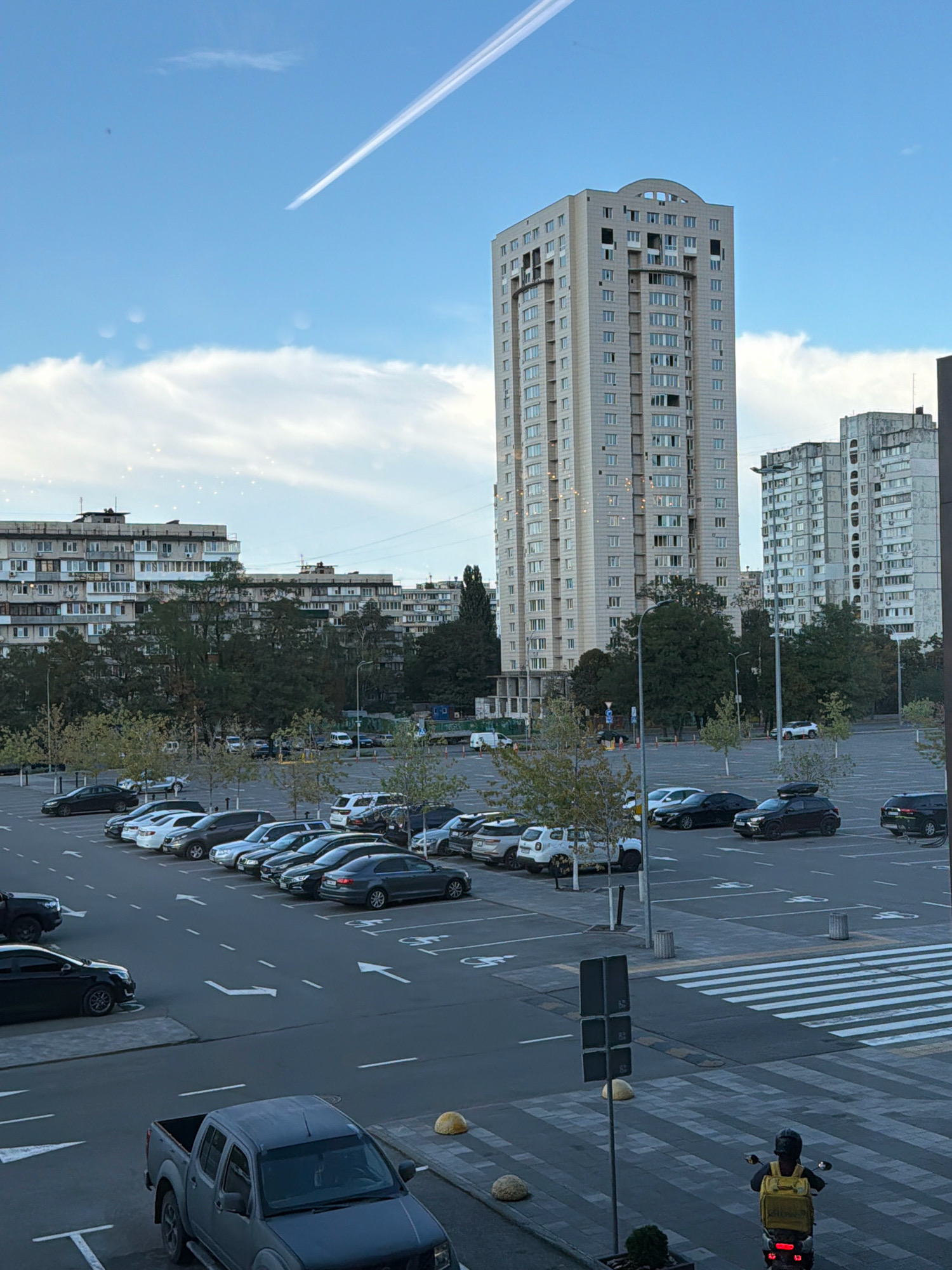 The image depicts a daytime scene in Kyiv, Ukraine, likely from an elevated vantage point, overlooking a large parking lot filled with various vehicles. In the background, several multi-story apartment buildings and a taller, modern high-rise are visible. The sky is blue with scattered white clouds, and a contrail from an aircraft stretches across it.
The parking lot occupies the foreground and mid-ground. Numerous cars, SUVs, and a pickup truck are parked or driving within the lot. Traffic lanes and parking spaces are marked with white lines, and some spaces are designated for disabled parking. A pedestrian crossing with striped markings is visible in the lower right portion of the image.
A person riding a scooter with a yellow delivery bag on the back is proceeding across the pedestrian crossing. The overall environment suggests an urban setting with residential and commercial buildings and infrastructure.