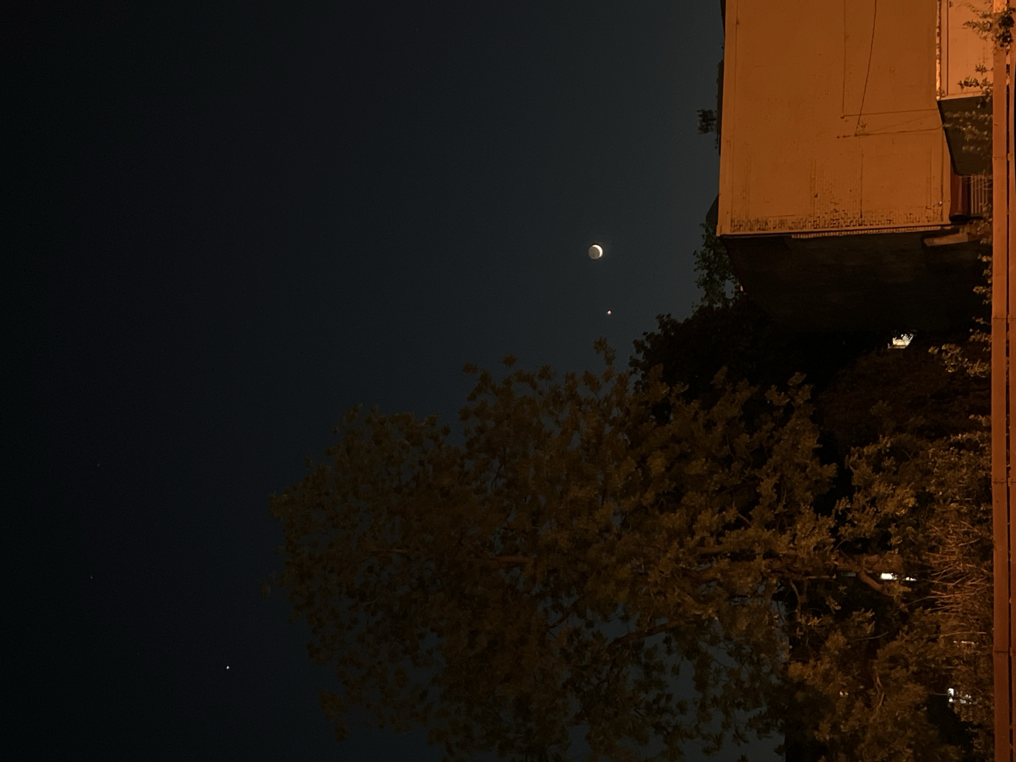 The image, taken at night, shows a crescent moon and a reddish planet (likely Mars) visible in the sky. A large tree with golden-lit leaves dominates the lower portion of the image, its branches reaching upwards. A building is positioned at the upper right, illuminated by a yellowish light, with a visible balcony. A thin vertical pole is to the right of the tree and building. The sky is a deep blue. The photograph captures a quiet nocturnal scene. No people are visible. The location is identified as City of Newburgh, United States of America.