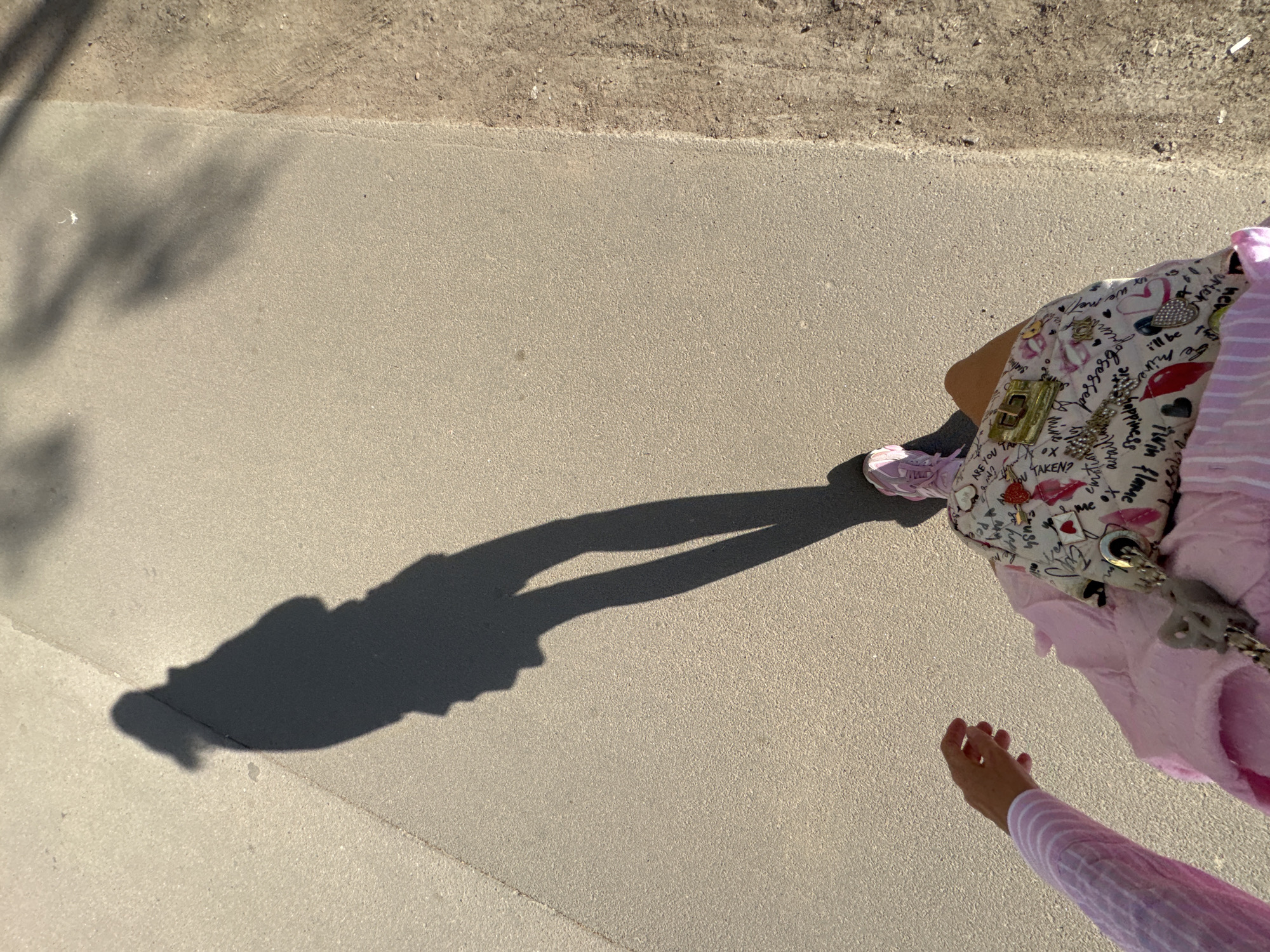The image shows a person walking on a paved surface, likely a sidewalk, with a sandy area visible to the side. The person is wearing pink sneakers and a pink and white striped long-sleeved shirt. They are carrying a white purse decorated with illustrations and text. The person's shadow is cast on the pavement in front of them.