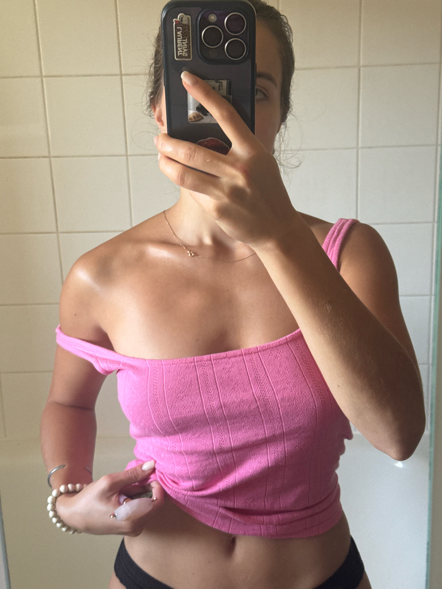 The image shows a young woman taking a mirror selfie in a bathroom. She is wearing a pink off-the-shoulder top and black underwear. She has a necklace on and bracelets on her left wrist. She is holding a rose quartz facial roller in her right hand. The bathroom has white tiled walls. The image does not contain any information about the location being in Vienne, France.