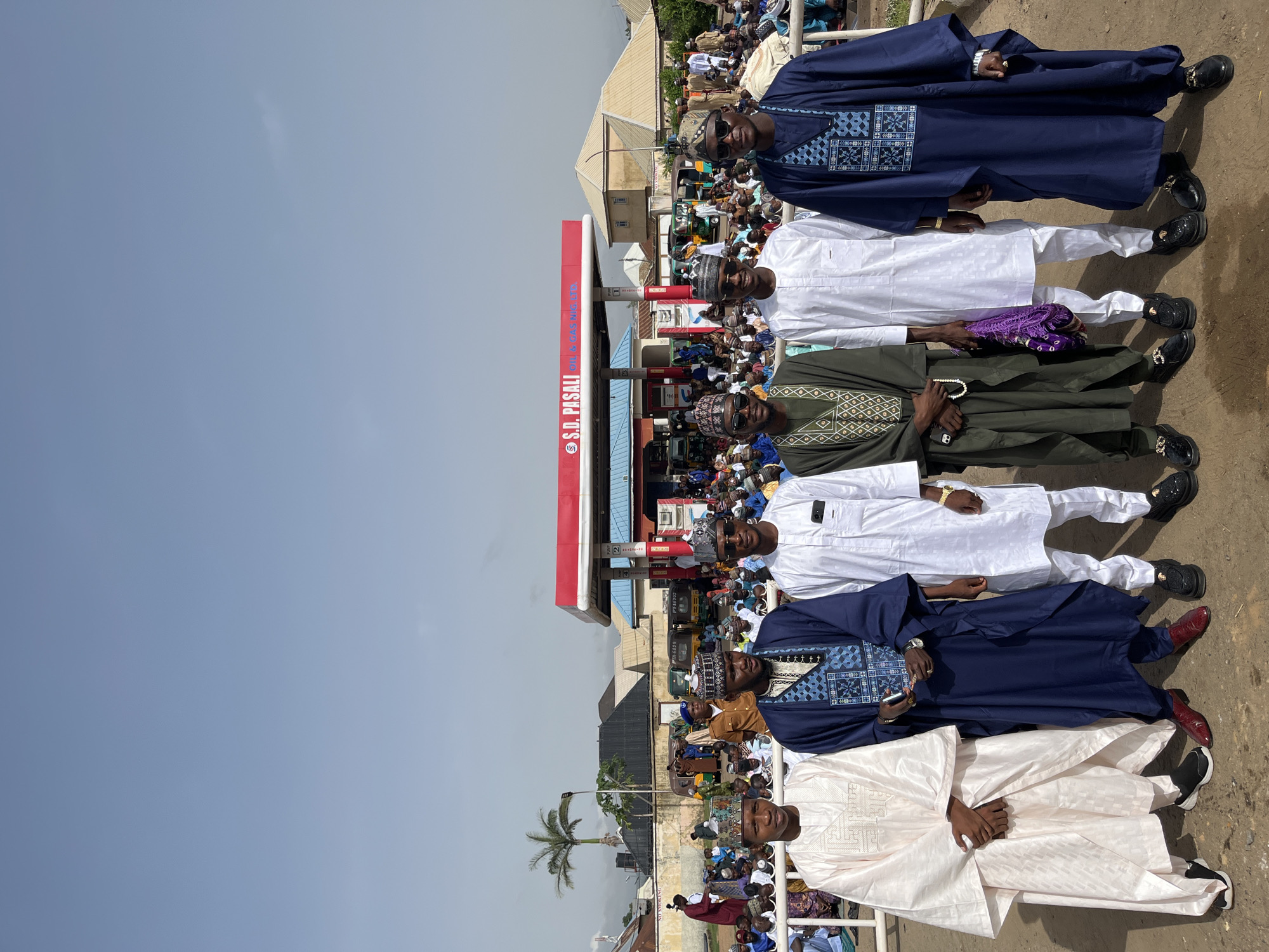 The image shows a group of six men standing in a row outdoors, likely in Jos, Nigeria, as indicated by the S.D. PASALI Oil & Gas station visible behind them. The men are wearing traditional African attire, including flowing robes, head coverings, and sunglasses. Behind the men, there is a large crowd of people, palm trees, and buildings. The sky is clear and blue. The scene appears to be captured during the day.