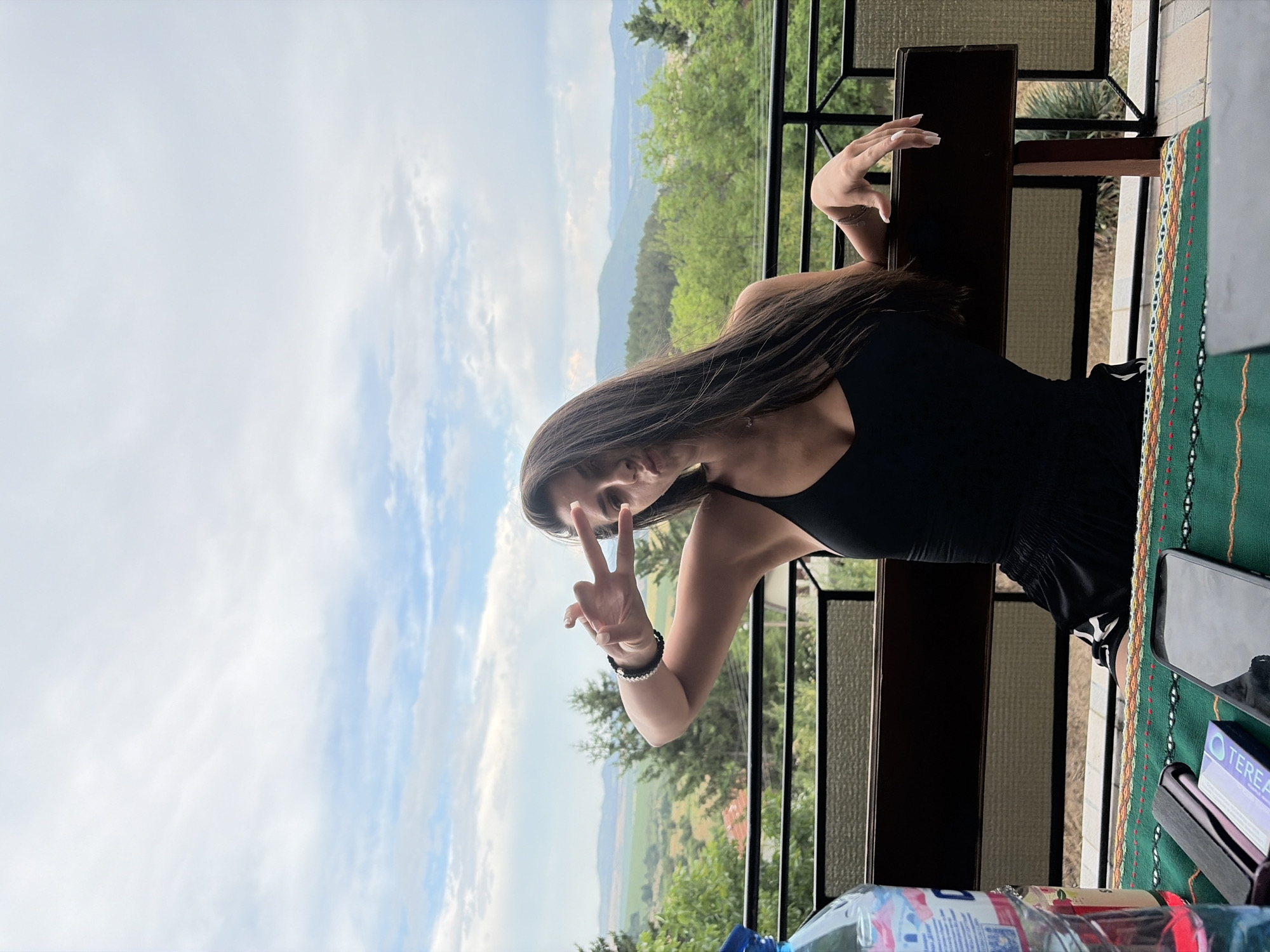 A young woman with long, dark hair is seated on a wooden bench on a balcony, outdoors. She is wearing a black tank top and black shorts. She is making a peace sign with her right hand, holding it near her face. A bottle of water and a smartphone are visible on a table in front of her, covered with a colorful textile. The background shows a scenic view of rolling hills, trees, and a cloudy sky, consistent with the landscape around Vladislavtsi, Bulgaria. The railing of the balcony is visible in the foreground and mid-ground.