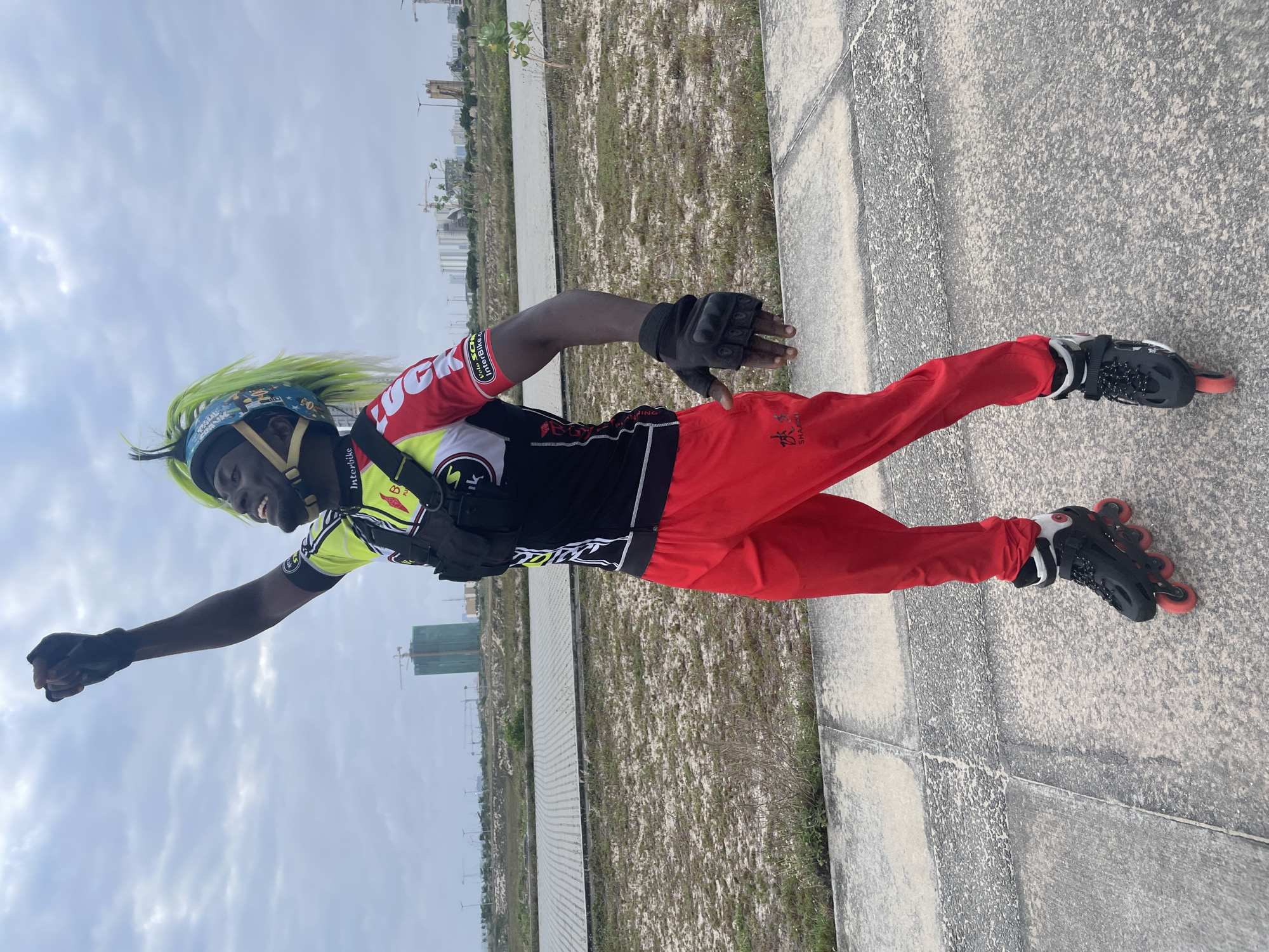 A man with bright green hair, wearing a helmet, a black and yellow and red cycling jersey with a chest pack, red pants, black gloves, and black rollerblades, stands on a concrete path in a grassy, undeveloped area. He is raising his right fist in the air. In the background, several tall buildings and construction cranes are visible against a cloudy sky, suggesting an urban or suburban area undergoing development. This scene is consistent with areas in the city of Lagos, Nigeria, characterized by ongoing construction and modernization.