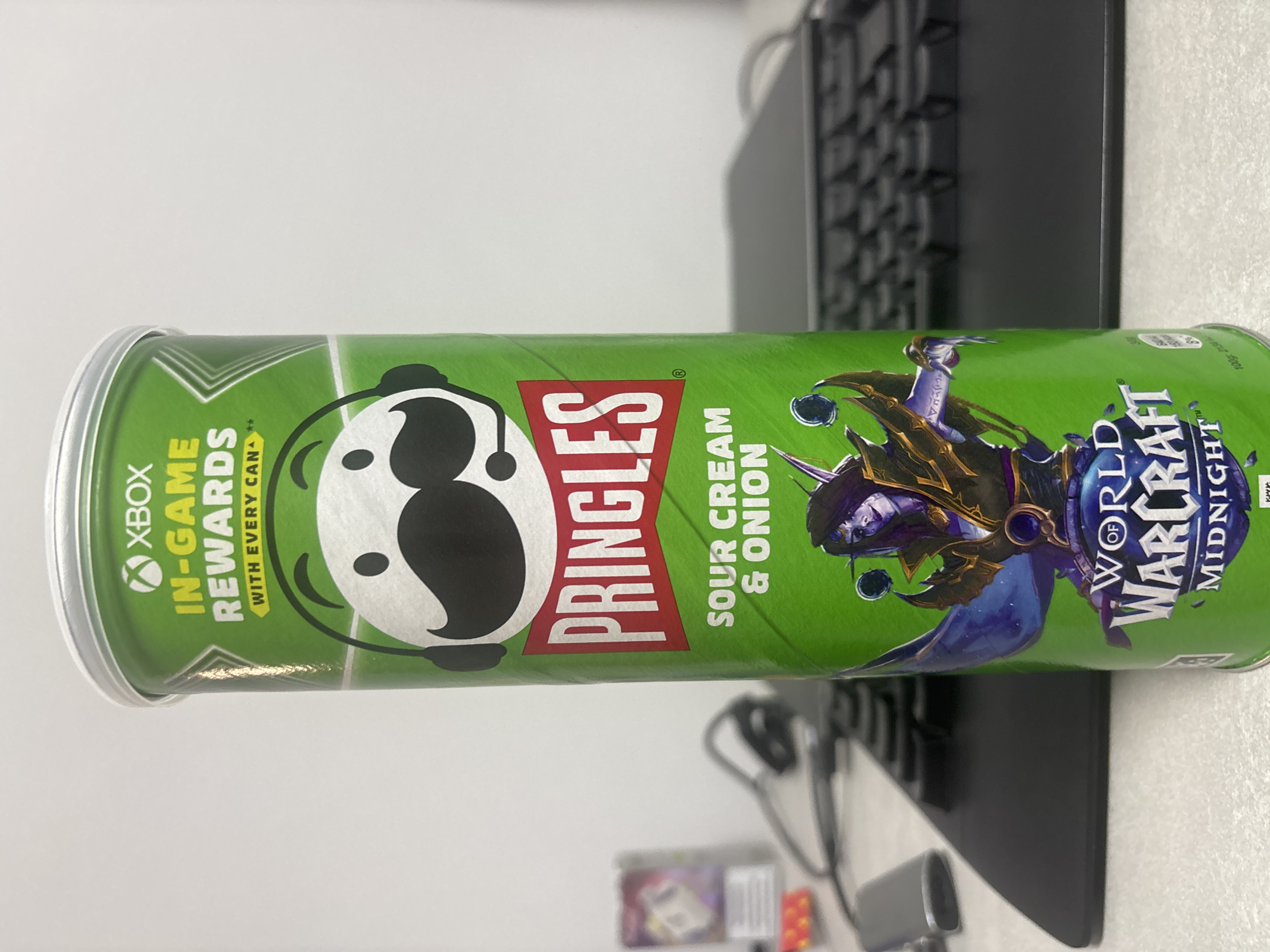 The image shows a can of Pringles Sour Cream & Onion chips, branded with Xbox in-game rewards and a World of Warcraft Midnight theme. The can is green, silver at the ends, and features the Pringles logo. It is positioned on a surface next to a black keyboard. No people are visible in the image. The location in Plovdiv, Bulgaria is unknown and not evident from the provided image.