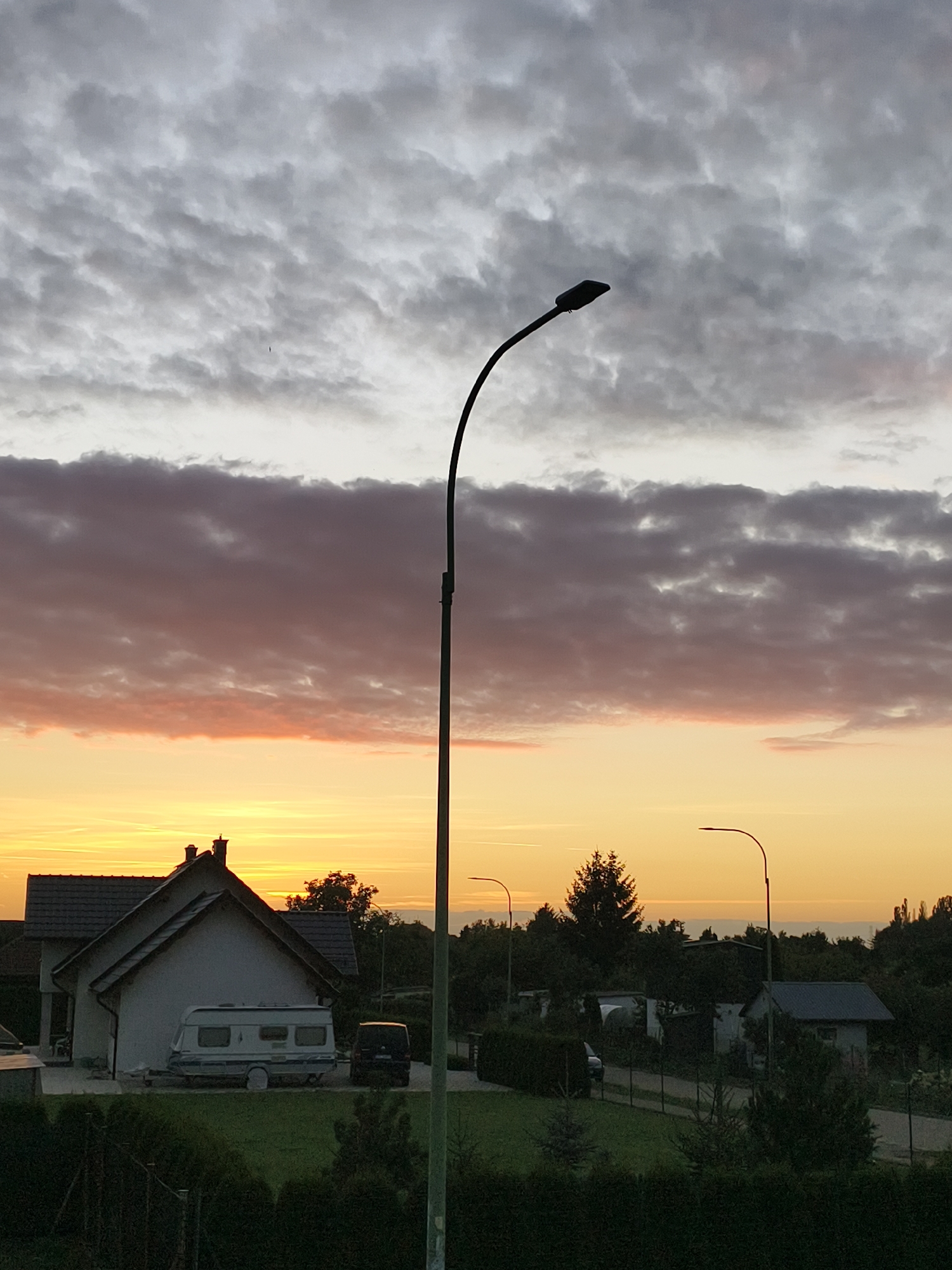 The image depicts a rural or suburban landscape at dusk, characterized by a dramatic sky filled with clouds in shades of gray, pink, and orange. A prominent street lamp with a curved pole stands in the foreground, its dark silhouette contrasting with the vibrant sky. In the middle ground, a house with a dark roof and a white facade is visible, with a caravan parked in front of it and a dark van to its right. The surrounding area features trees, smaller buildings, and a neatly trimmed hedge in the foreground. The horizon is dotted with more trees and structures, suggesting a sparsely populated area. The overall impression is one of a quiet evening scene, possibly in the outskirts of Tczew, Poland, given the reference to the city. There are no people or animals visible in the frame.