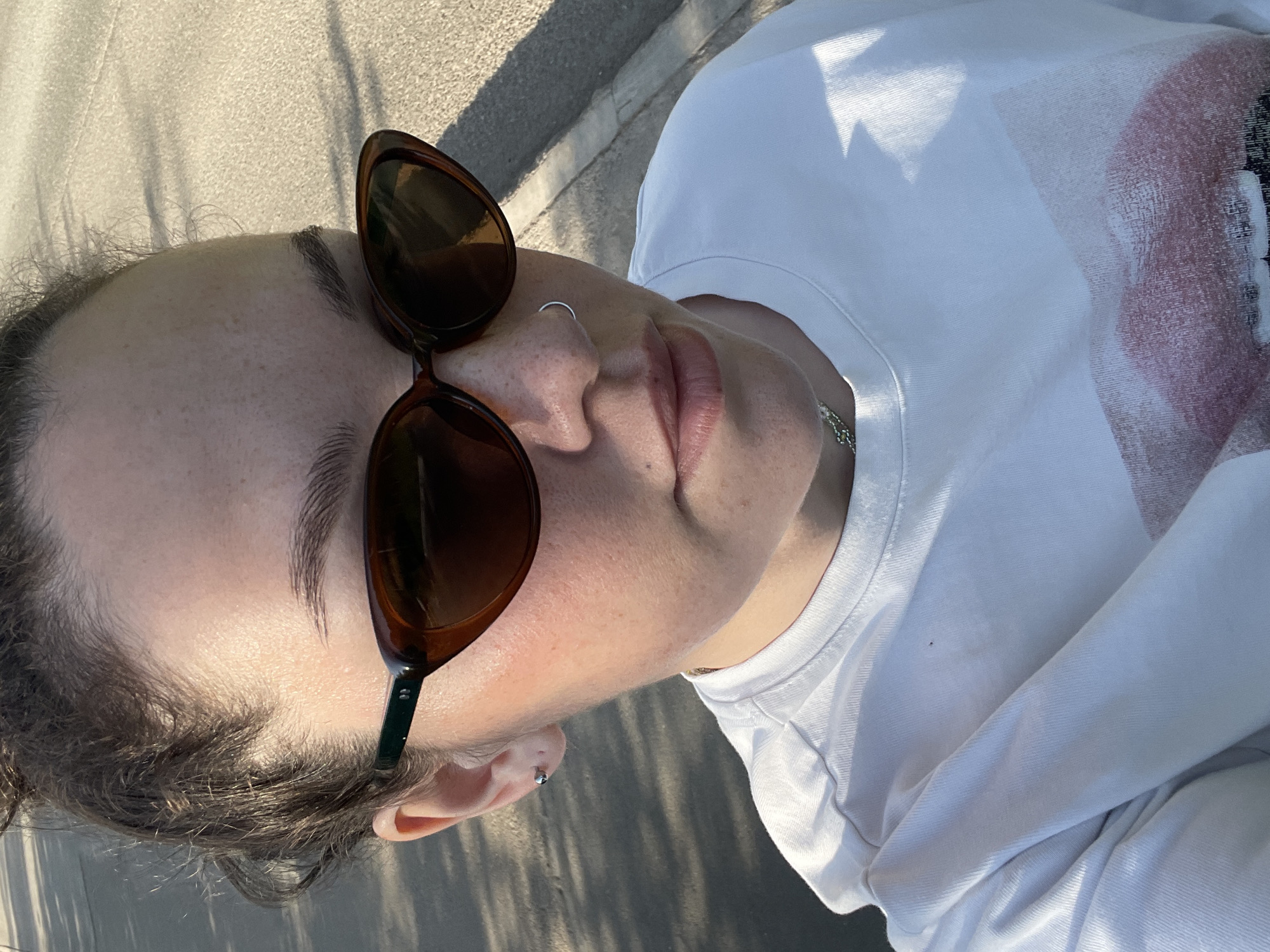 A close-up, canted angle shot features a person wearing brown-framed sunglasses. A silver nose ring is visible in the left nostril, and a silver stud earring is in the left ear. The person is wearing a white T-shirt with a design on the right side. The background consists of an indistinct gray surface, possibly a sidewalk. There is no information that supports the location of this image in Izhevsk, Russia.