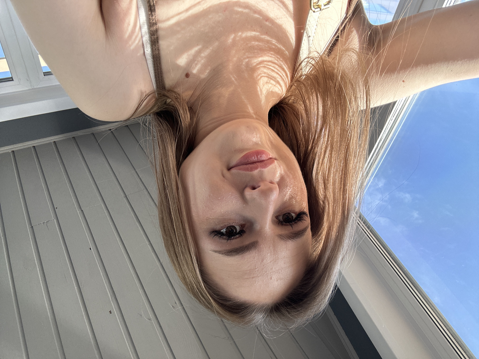 A young woman is captured in an inverted close-up selfie, with her face centered at the bottom of the frame. She looks directly at the camera with a neutral to slightly smiling expression, her lips slightly parted. Her long, light brown hair frames her face and shoulders. She wears a light-colored top, with a strap visible on her right. Strong sunlight illuminates her, casting distinct, organic shadow patterns across her chest and neck, indicative of natural light filtering through foliage or a patterned screen.
Above her, through what appears to be a window or opening, a clear blue sky with a few wispy white clouds is visible, suggesting a sunny day. Below the window, a light grey surface with horizontal paneling or slats is present, possibly part of a wall or railing. The image emphasizes the interplay of light and shadow on her skin and the bright outdoor setting. No discernible text is visible.
