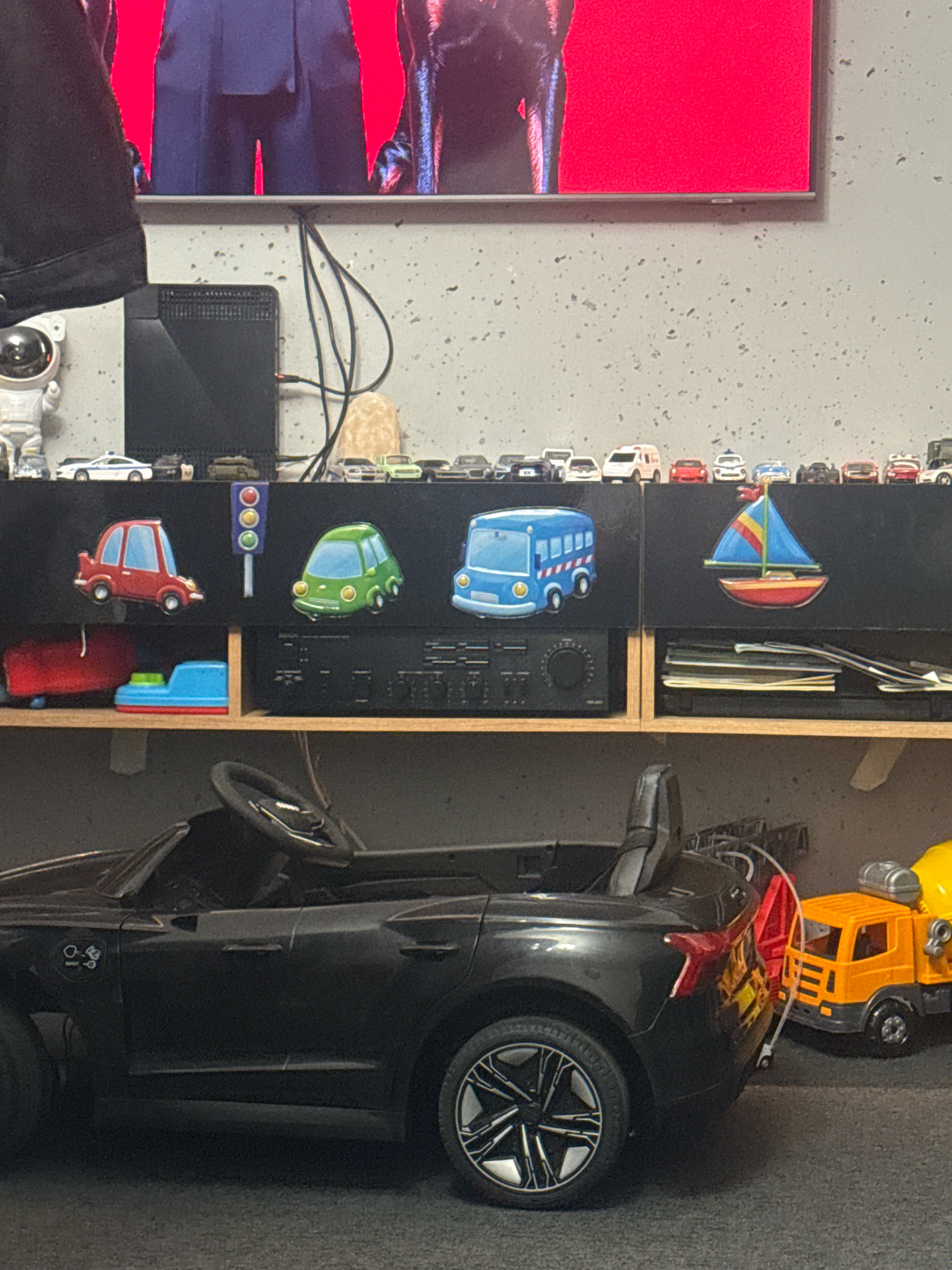 The image depicts a collection of toy vehicles displayed on shelves. In the foreground, a large black toy car is visible, its interior with a steering wheel and dashboard partially seen. To the right of the black toy car, a smaller orange toy construction vehicle with a cement mixer is present. The middle shelf displays decorative decals of a red car, a green car, a blue bus, and a sailboat. Above these decals, a row of miniature toy cars and vehicles are lined up. The background shows a television screen displaying a red background with people. The overall environment suggests a room or storage area. The location of Gorzow, Poland is not visually identifiable within the image.