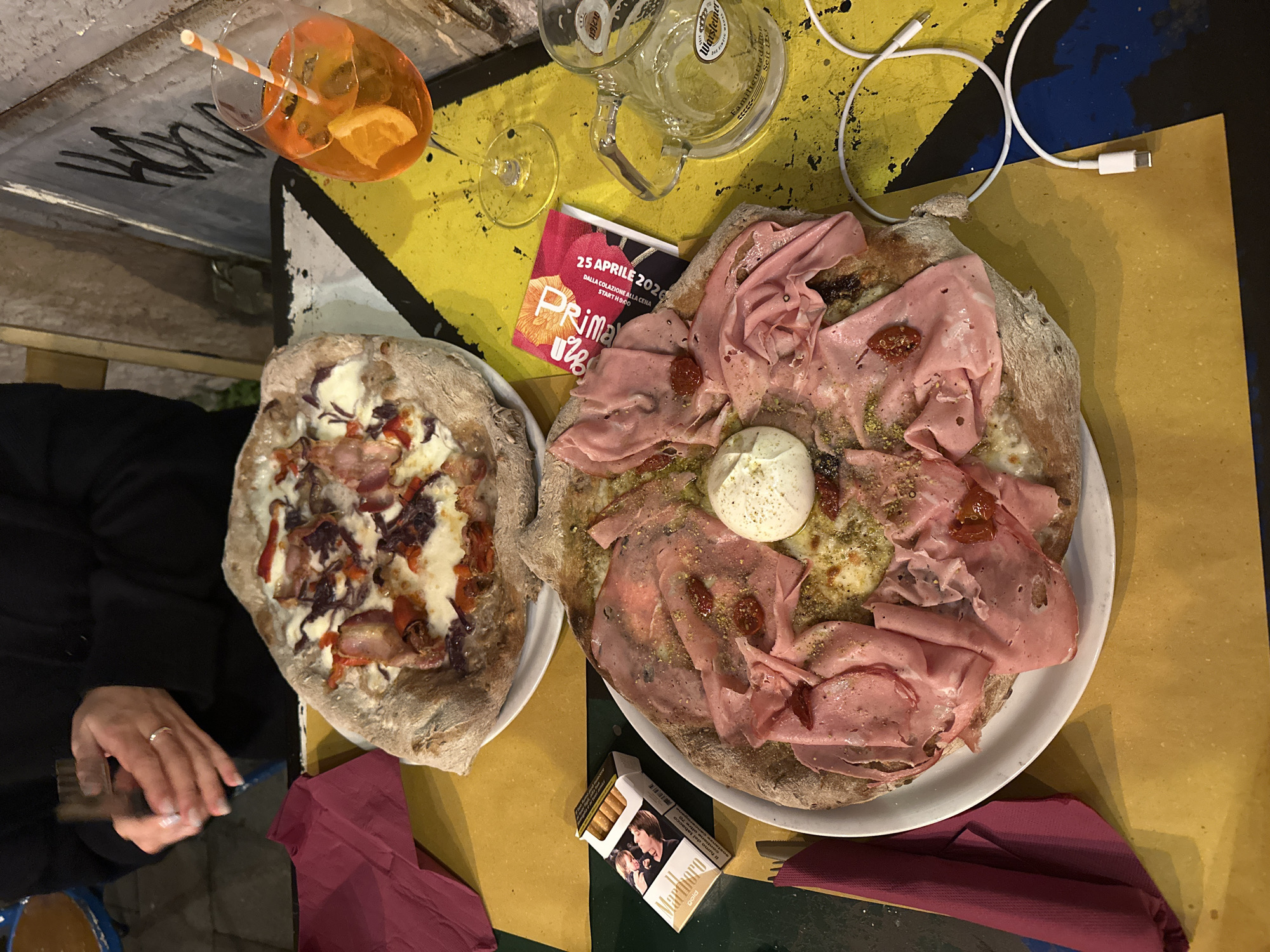 The image shows a high-angle view of a table scene in Catania, Italy, set with a variety of food and beverage items. Two pizzas are centrally placed: one is topped with cheese and red vegetables, and the other is covered with thin slices of meat, a white ball in the center, and smaller red pieces. A promotional card with the text "25 APRILE 2026" is visible near a glass mug and a wine glass. An orange cocktail with a straw is located in the upper left. A pack of Marlboro Gold cigarettes lies near the pizza topped with meat. A white USB charging cable is also visible. In the lower left corner, part of a person's arm and hand holding a black object are seen. The table's surface is primarily yellow with a dark green and blue geometric pattern.