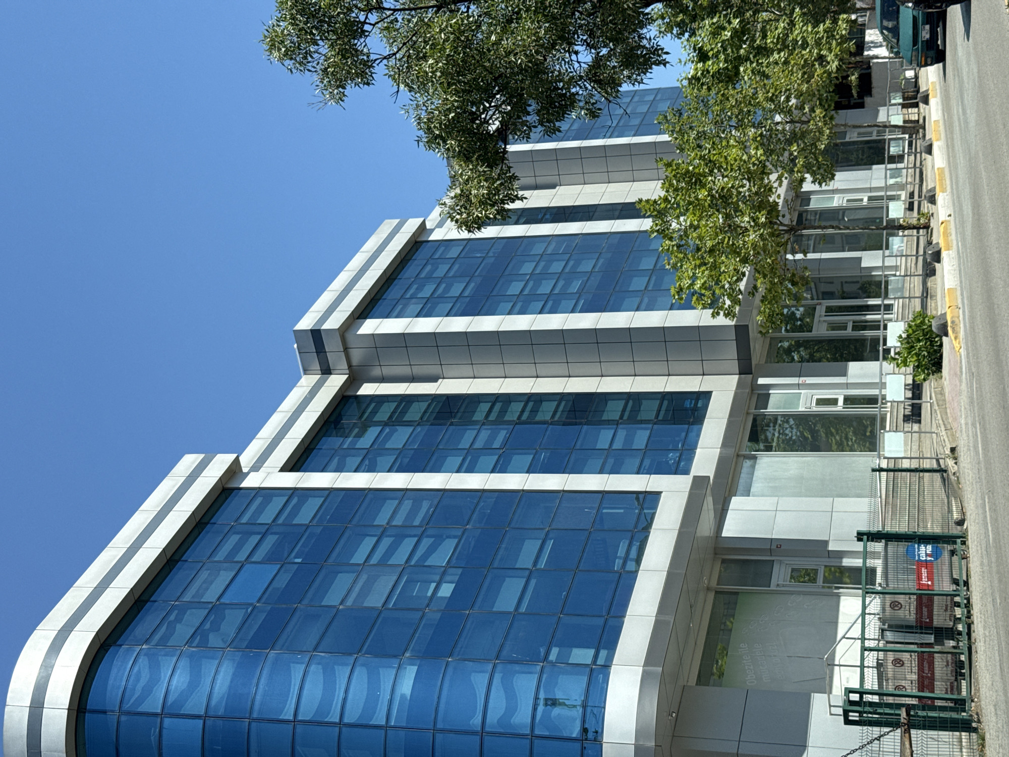 A modern, multi-story office building with a curved facade and predominantly blue glass windows stands under a clear blue sky. The building's exterior is a combination of brushed metal panels and white trim. In the foreground, a paved road runs along the front of the building, with a sidewalk and some trees. A green metal fence encloses an area in front of the building, where several large containers or bins are visible. A dark green car is parked on the right side of the road. Some green foliage from trees can be seen partially obscuring the building. The overall scene depicts a typical urban or suburban commercial environment in Sancaktepe, Turkey.