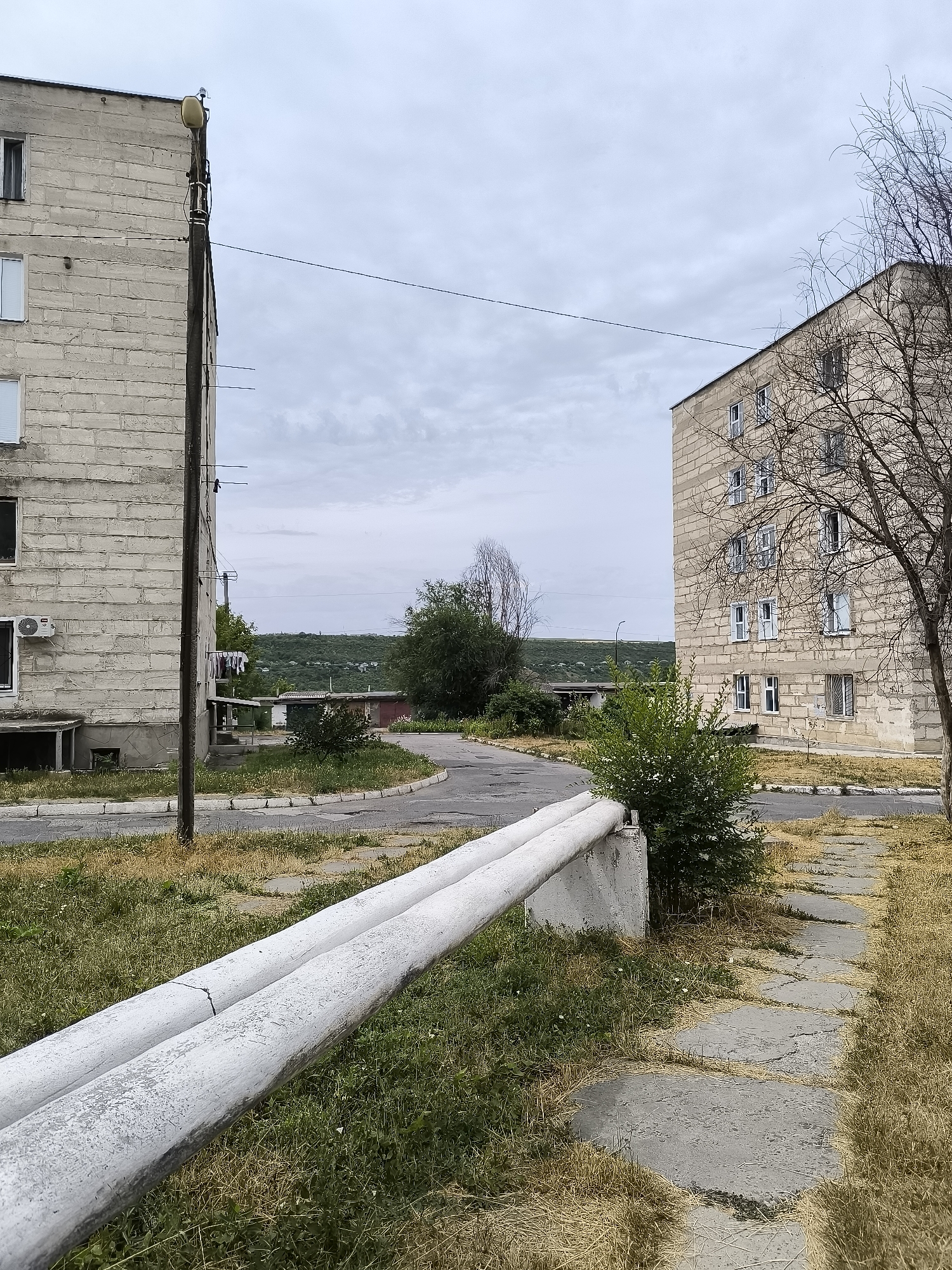 The image shows an exterior scene in the city of Rybnitsa, Moldova, featuring two large, multi-story concrete apartment buildings under an overcast sky. In the foreground, a pair of large, white-painted pipes run horizontally across the frame, supported by a concrete structure. These pipes appear to be part of an above-ground utility system.
To the left, a portion of a light-colored, block-constructed apartment building is visible. It has multiple windows, some with blinds. A concrete utility pole stands in front of this building, with various wires and what appears to be laundry hanging from a line. An air conditioning unit is mounted on the side of the building.
To the right, another similar apartment building is visible, also constructed from concrete blocks, with numerous windows. A bare tree with intricate branches is positioned in front of this building, its silhouette stark against the sky.
The ground between the buildings is a mix of cracked concrete slabs and overgrown dry grass, indicative of an unkempt urban environment. A paved road or pathway curves around the buildings. In the background, a hilly, vegetated landscape is visible under the diffused light of the cloudy sky, with some smaller structures and trees dotting the terrain. The overall impression is one of an older, possibly underdeveloped, residential area.
