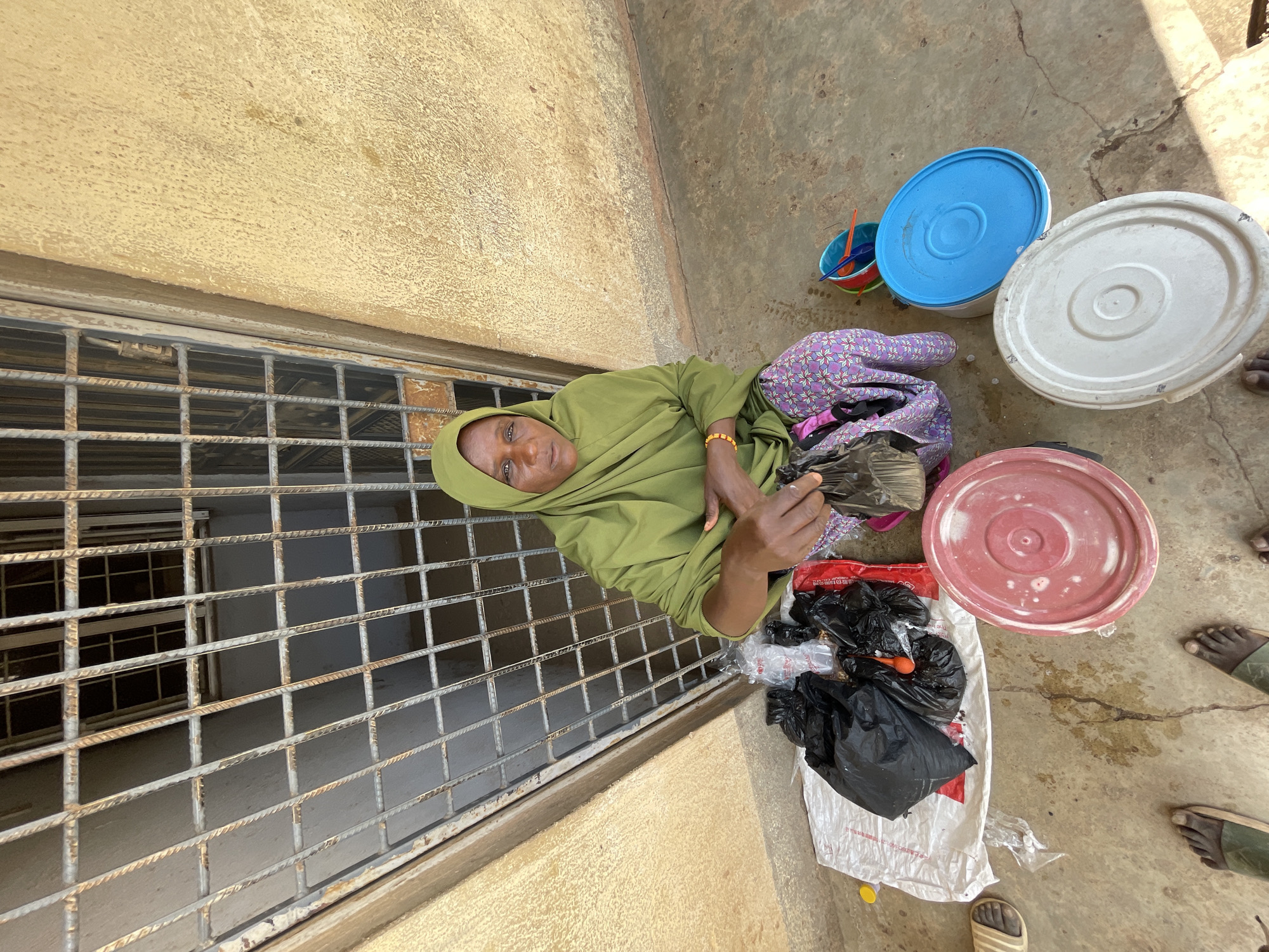 A medium-shot, full-body view looking down at an angle, reveals a woman in Jos, Nigeria. She sits on the ground near a barred window, wearing a light green hijab and a patterned purple skirt. Her right hand is extended, holding a dark, unidentifiable object, while her left hand rests in her lap.
The woman is surrounded by various objects. To her right, are several plastic buckets with lids, stacked near black plastic bags on top of a white shopping bag. Near the edge of the frame, portions of feet are visible, hinting at the presence of other individuals nearby. The ground appears to be a concrete surface, likely an outdoor or semi-outdoor setting. The window behind the woman is barred with a metal grate, suggesting an institutional or commercial environment. The wall adjacent to the window is a rough, light brown color.