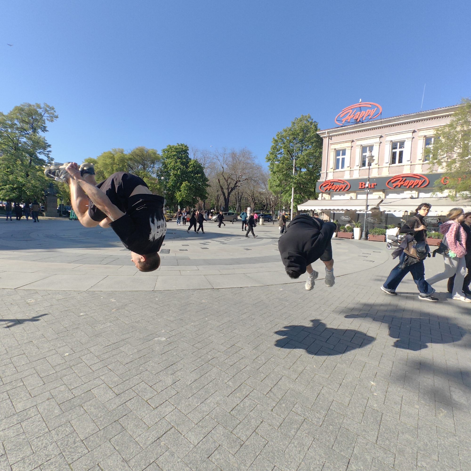 The image depicts two individuals performing acrobatic flips in a public square. The square appears to be the central pedestrian zone in Varna, Bulgaria, judging by the recognizable building of the "Happy Bar & Grill" restaurant chain in the background.
The person on the left is upside down and wearing a black shirt with white markings and dark shorts, performing a backward flip. The person on the right is also upside down and wearing a black hoodie and shorts, also performing a flip. Shadows cast on the cobblestone ground suggest bright, sunny conditions. Several other individuals are walking through the square, including some near the restaurant. Trees line the perimeter of the square. The sky is clear and blue.