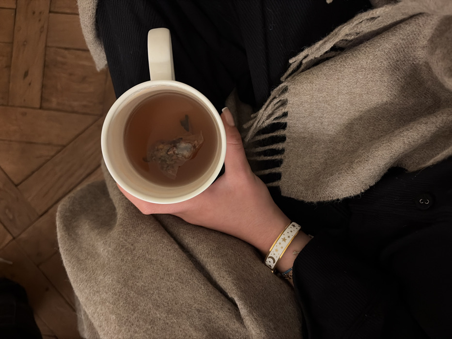 A right hand holds a white ceramic mug containing an amber liquid and a steeping tea bag. The hand features manicured nails with light polish, and a white and gold-toned bangle bracelet with moon and star motifs is worn on the wrist. The individual is partially clad in a dark, likely black, long-sleeved garment, and is covered by a light brown, fringed blanket or throw. The background reveals a light brown wooden floor with a herringbone pattern, typical of traditional Parisian interior architecture. The scene depicts an indoor domestic setting, consistent with an apartment in Paris, France.
