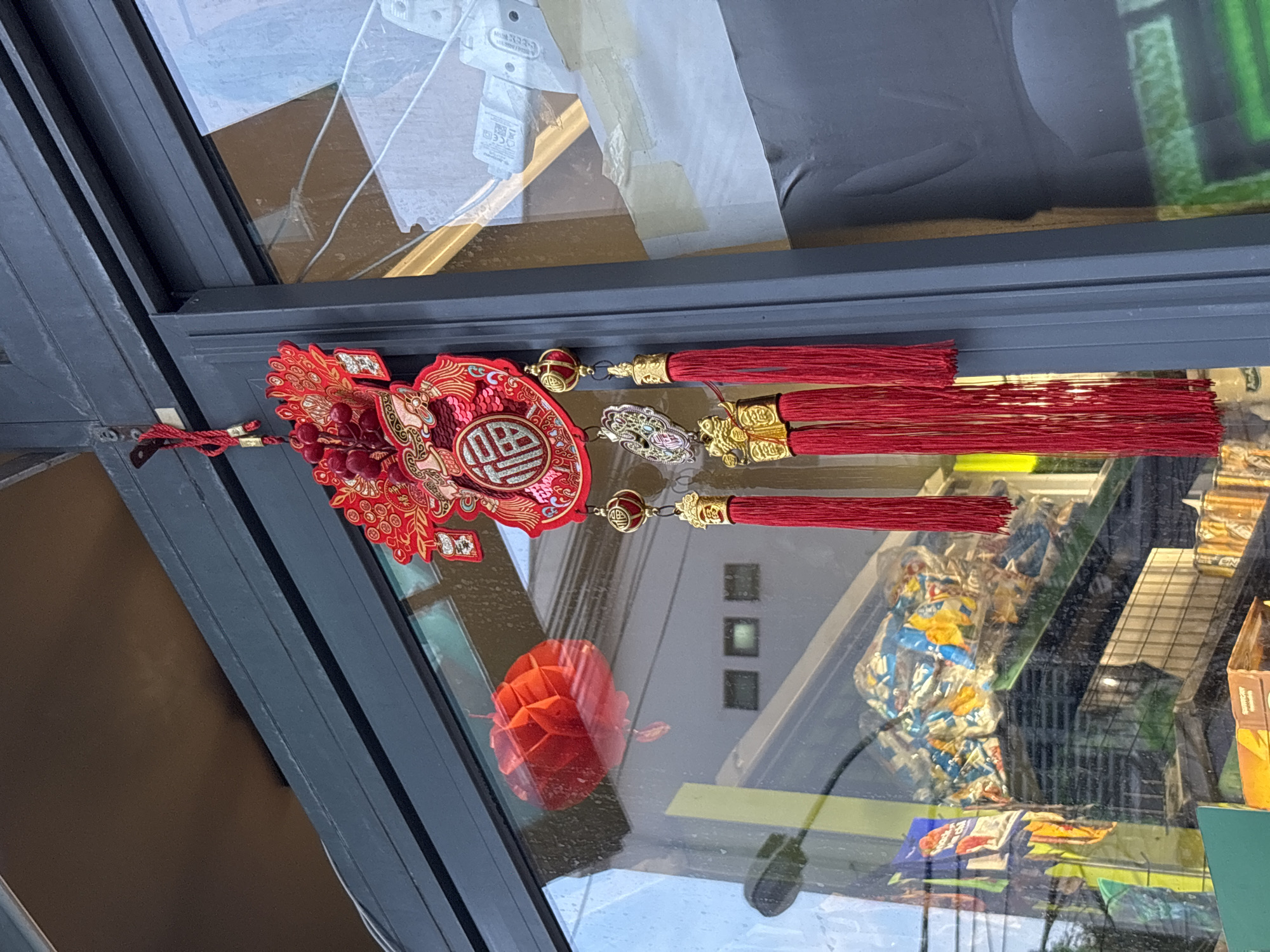 A red and gold Chinese New Year decoration with tassels hangs from a metal fixture on a dark gray, sliding glass door frame in Nha Trang, Vietnam. The decoration features a central circular emblem with Chinese characters, surrounded by intricate red and gold filigree. A red paper lantern is reflected in the glass behind the door. Inside the store, beyond the glass, shelves are visible filled with various goods, including bags of snacks, canned drinks, and other items. The overall environment suggests a commercial establishment, likely a shop or market, decorated for a festive occasion.