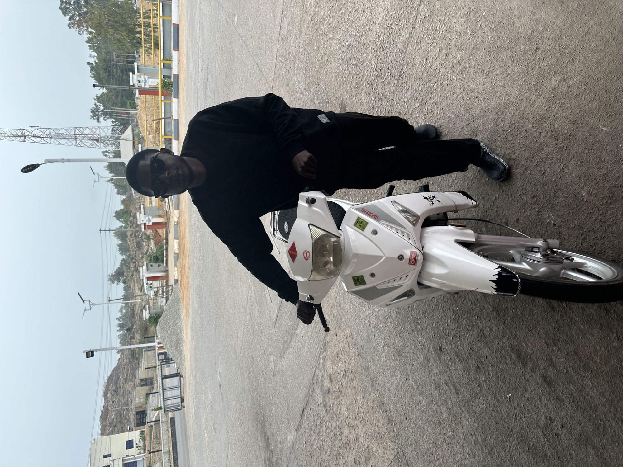 A man in dark sunglasses and black clothing lies prone on a white moped. The moped is parked on gray asphalt. In the background are utility poles, buildings, a communication tower, trees, and a pile of gravel. The location is Jos, Nigeria.