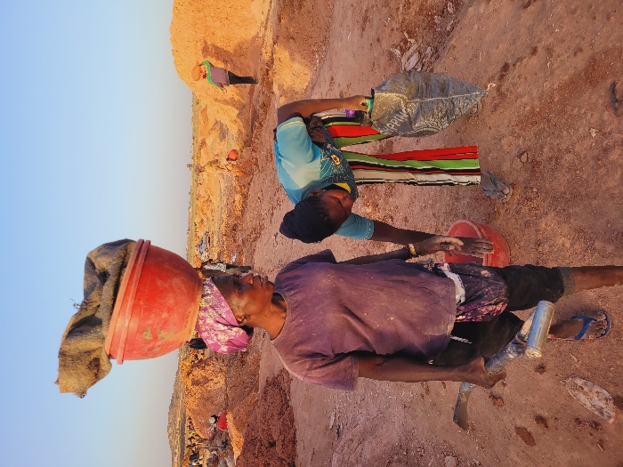 This image depicts an outdoor scene in the city of Shen, Nigeria, at what appears to be a manual excavation or mining site under a clear, pale blue sky. The ground is reddish-brown, dry, and dusty, with numerous large mounds of earth visible across the wide landscape.
In the foreground, two adult females are engaged in labor. The woman on the left, with dark skin, wears a dusty light purple t-shirt, dark trousers, blue flip-flops, and a patterned pink and purple headscarf. She balances a large, red plastic basin containing a brown burlap sack on her head. In her right hand, she holds a hoe-like tool with a metallic handle and a silver cylindrical object, possibly a can or bottle. Her gaze is directed towards the right side of the frame.
To her right, slightly behind and bent forward, is another adult female with dark skin. She wears a light blue short-sleeved top with a faint design, brightly colored vertically striped pants (red, green, yellow, black), and grey flip-flops. Her left hand holds a black plastic bag, partially displaying the word "SHOPPING," while her right hand is extended towards a red plastic bucket on the ground.
In the mid-ground and background, additional indistinct figures are seen scattered among the earth mounds, also appearing to be engaged in similar manual tasks. One figure on the far right carries a red basin on their head. A white vehicle is partially visible further back among the mounds.