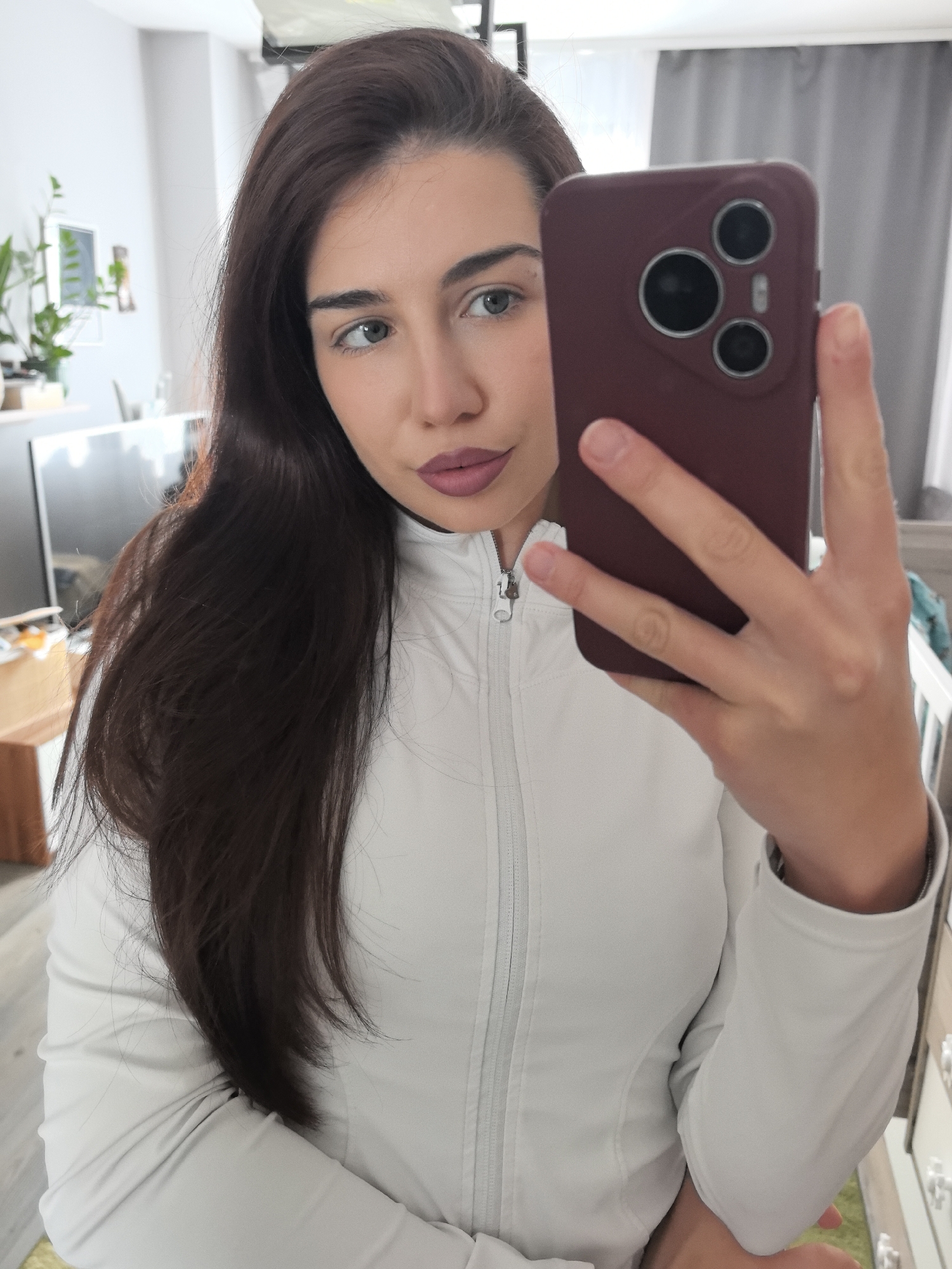A young woman with long, dark brown hair and green eyes is taking a mirror selfie indoors. She is wearing a light-colored, possibly white or light grey, long-sleeved athletic top with a zipped collar. Her lips are painted with a muted reddish-purple lipstick, and her gaze is directed slightly to her left. She holds a smartphone with a dark red or burgundy case in her right hand, obscuring part of her face. The phone case features three circular camera cutouts on its back.
The background suggests a residential interior, consistent with a home in Stara Zagora, Bulgaria. To the left, a potted plant, a framed picture, and a television screen are partially visible against a light-colored wall. To the right, a window with light grey curtains and a piece of white furniture (possibly a chest of drawers or a crib) can be seen. The flooring appears to be light-toned. The lighting indicates it is daytime. The woman's expression is neutral with a slight pout. No visible text is present in the image.