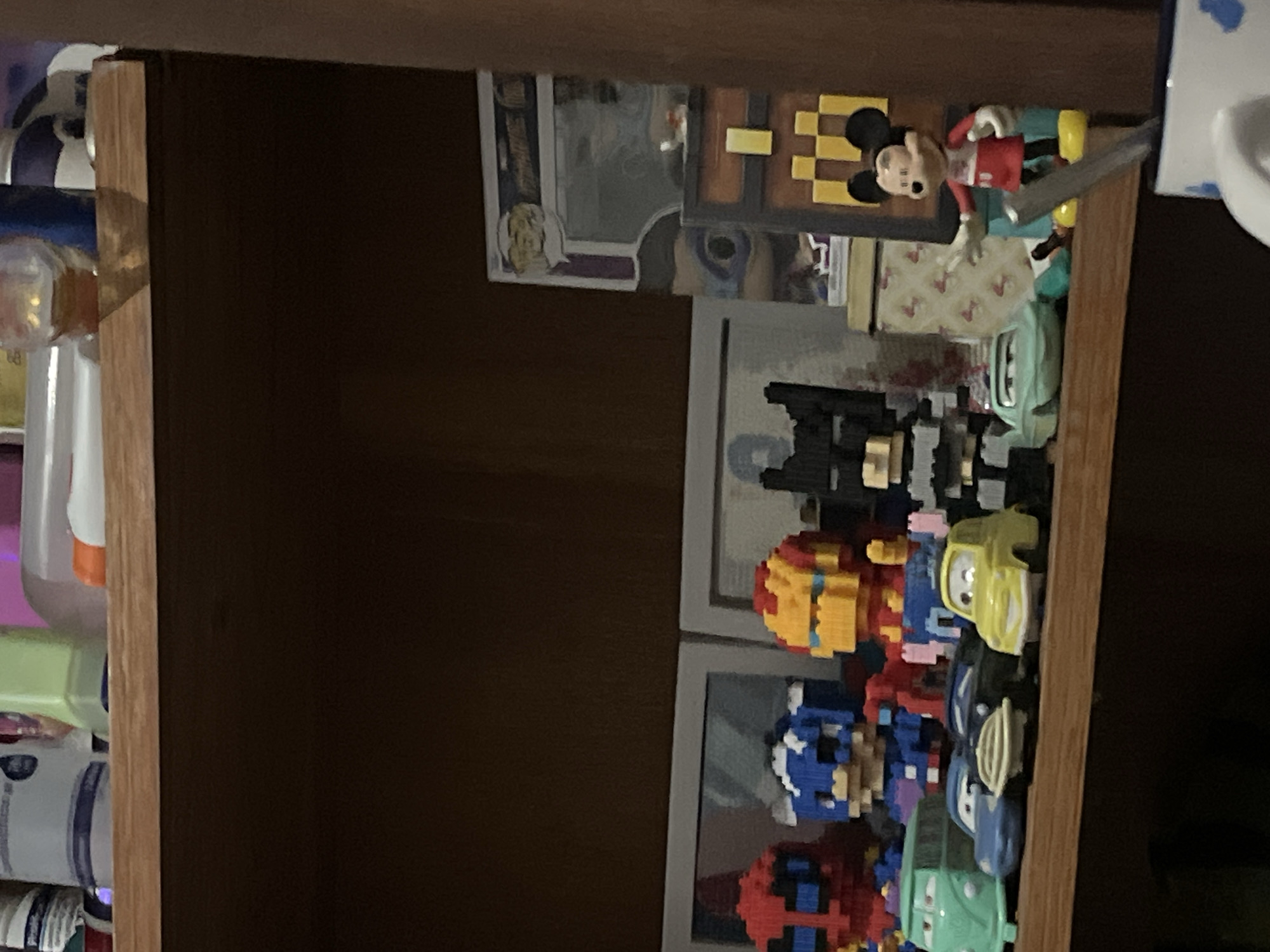 A shelf is lined with toys and other items. On the shelf, from left to right, are: a red and blue toy figure that appears to be Spider-Man, a blue and white toy figure that appears to be Captain America, a yellow and red toy figure that appears to be Iron Man, a black toy figure that appears to be Batman, and a Mickey Mouse figure. In front of these figures are toy cars, including a light green van, a dark blue car, a black car with a white stripe, and a yellow car. Behind the figures is a framed picture or poster, and to the right of that is a wooden chest with pixelated designs. The background is dark, suggesting a dimly lit room. The items are arranged on a wooden shelf. The image is taken from a slightly low angle. The location is specified as Santa Ana, Venezuela.