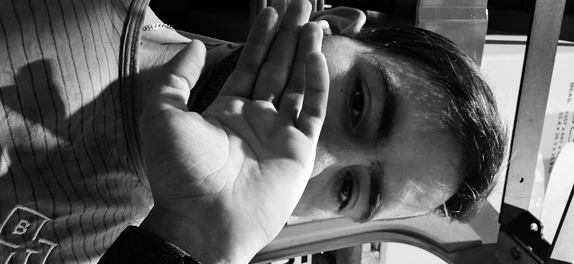 This black and white, close-up image features a person, likely a young adult, with their face partially obscured by their right hand. The image is presented with a horizontal orientation, though the subject appears to be leaning or the photo was taken at an angle, with their head positioned towards the right side of the frame and their torso on the left.
The person has dark hair and dark eyes, with one eye looking directly at the camera. Their right hand is held up, palm open and facing the viewer, covering the lower part of their face. The fingers are slightly splayed, revealing intricate lines and creases on the palm due to the strong, directional lighting.
The individual is wearing a striped tank top or sleeveless shirt. A silver-colored chain is visible around their neck. On the lower left side of the shirt, an embroidered or printed motif featuring a large letter 'B' and another letter, possibly 'T', below it, is visible.
The lighting is intense, casting stark shadows across the striped fabric of the shirt and emphasizing the contours of the hand and the visible parts of the face. This suggests a strong light source, possibly direct sunlight or a powerful artificial light.
In the upper right background, details of an indoor setting are discernible. A light-colored object, likely product packaging or a label, displays several pieces of text. The word "MEAS:" (measurements) is visible, followed by numerical dimensions like "1357 x 667" and "53.4 x 26.3 x 25.9". The partially visible word "IDEAS" further suggests this could be packaging for furniture or a similar item, potentially from a brand like IKEA. A dark, angular object is also present in the background.
The activity appears to be a casual portrait. The hand gesture could imply shyness, concealment, or a deliberate pose. The obscured face makes it difficult to discern a clear emotion, but the visible eye maintains direct contact with the camera. The image contains no explicit visual cues identifying the location as Plovdiv, Bulgaria.