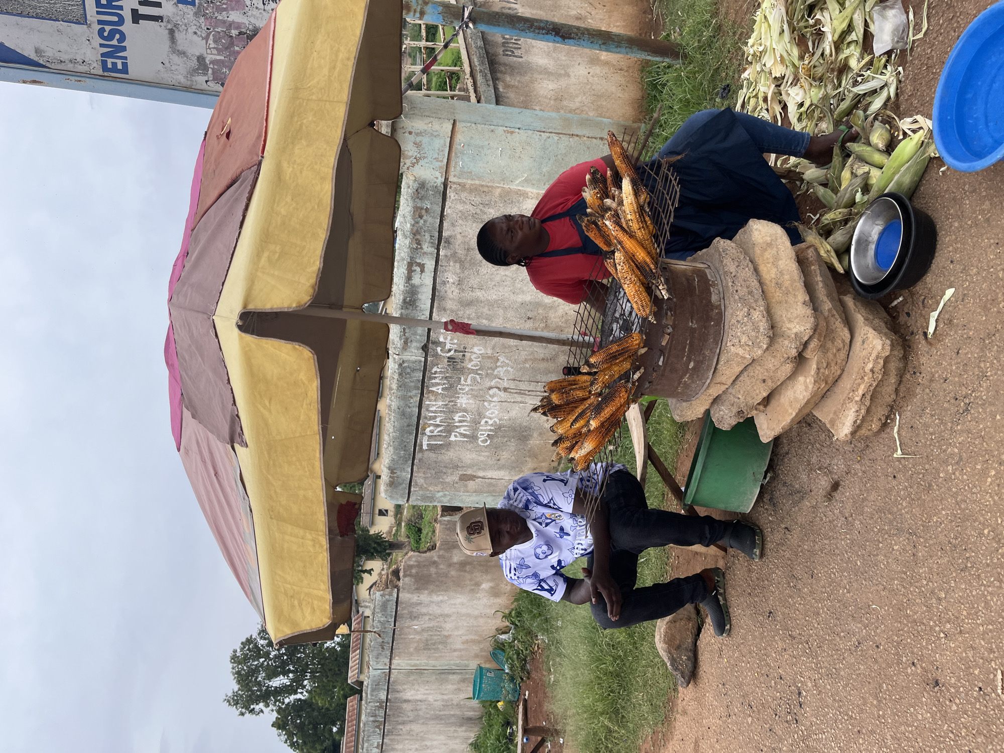 The image shows a street vendor selling roasted corn in Jos, Nigeria. Two men are present. One man, wearing a red shirt and a blue apron, is tending to the corn roasting on a grill. The other man, wearing a white and blue patterned shirt and a baseball cap, is sitting nearby.
The scene is set outdoors, with a large, colorful umbrella providing shade. A pile of corn husks and unhusked corn is visible, as well as cooking equipment including a metal grill, a large blue plastic basin, and a smaller metal bowl with a blue lid. Several large, rough stones are stacked near the grill, possibly used as a makeshift stove or base. The ground is a mixture of gravel and asphalt. In the background, a concrete wall with some graffiti and greenery can be seen.
The time of day appears to be daytime, likely overcast or cloudy, as indicated by the diffused light and the lack of strong shadows. The overall mood seems to be one of commerce and daily life.
Visible text on the wall behind the vendors reads: "TRAIN AND PAID 091309123". The context of "TRAIN AND PAID" is unclear without further information, but the numbers likely represent a phone number or transaction details.
