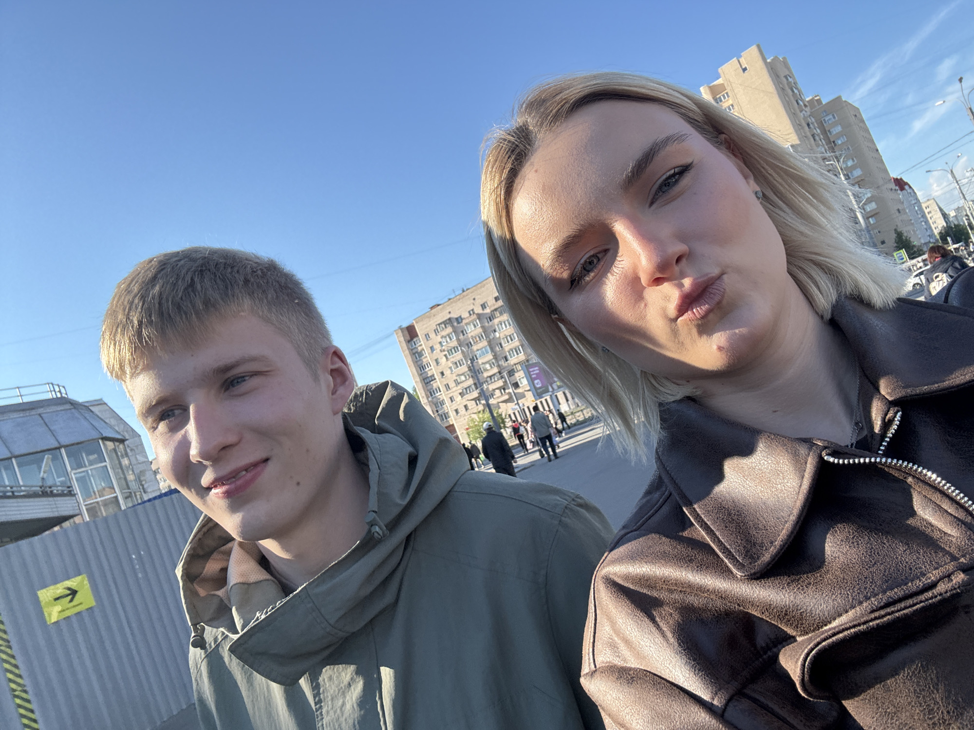 A selfie, taken outdoors in Vsevolozhsk, Russia, features two people. On the left is a young man with short, light brown hair, wearing a green hooded jacket. He is partially obscured by a gray construction barrier with a yellow directional arrow. To the right is a young woman with blonde hair, wearing a brown leather jacket and making a kissing face toward the camera. In the background, apartment buildings are visible under a bright blue sky. Several people walk along a sidewalk. Sunlight casts shadows and highlights across the scene.