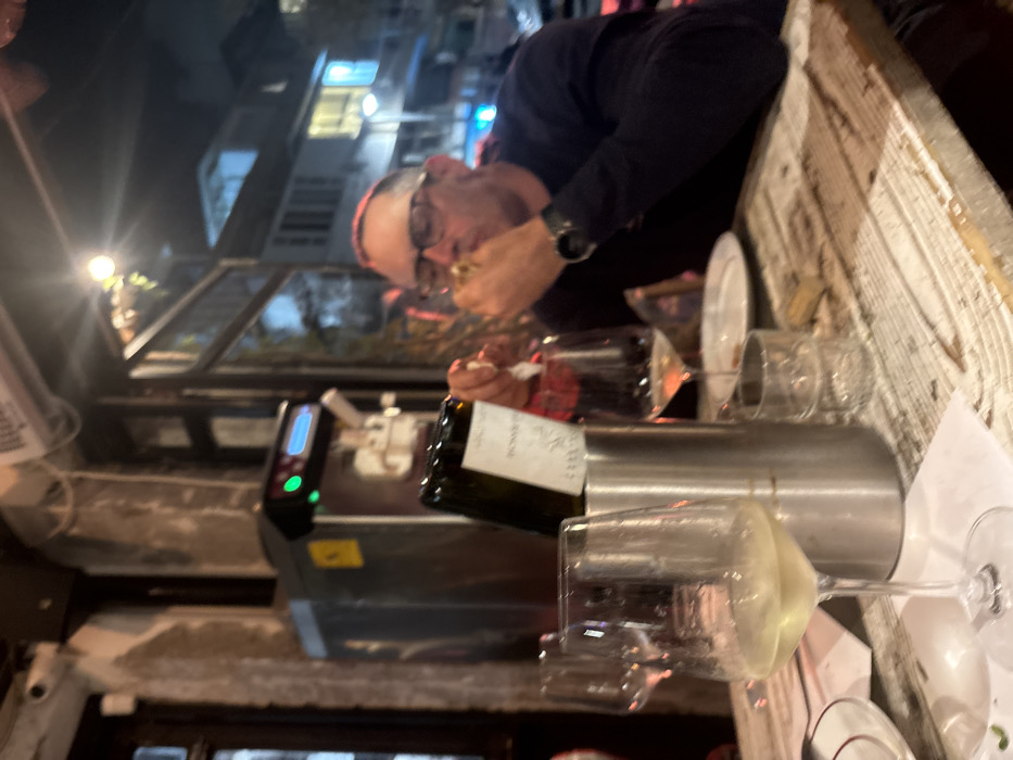 A man in a dark shirt and glasses eats while seated at a table with multiple wine glasses, a wine bottle, and a chiller. A machine with a digital display sits nearby. Overhead, a glass roof reveals a cityscape at night. The table is aged and wooden.