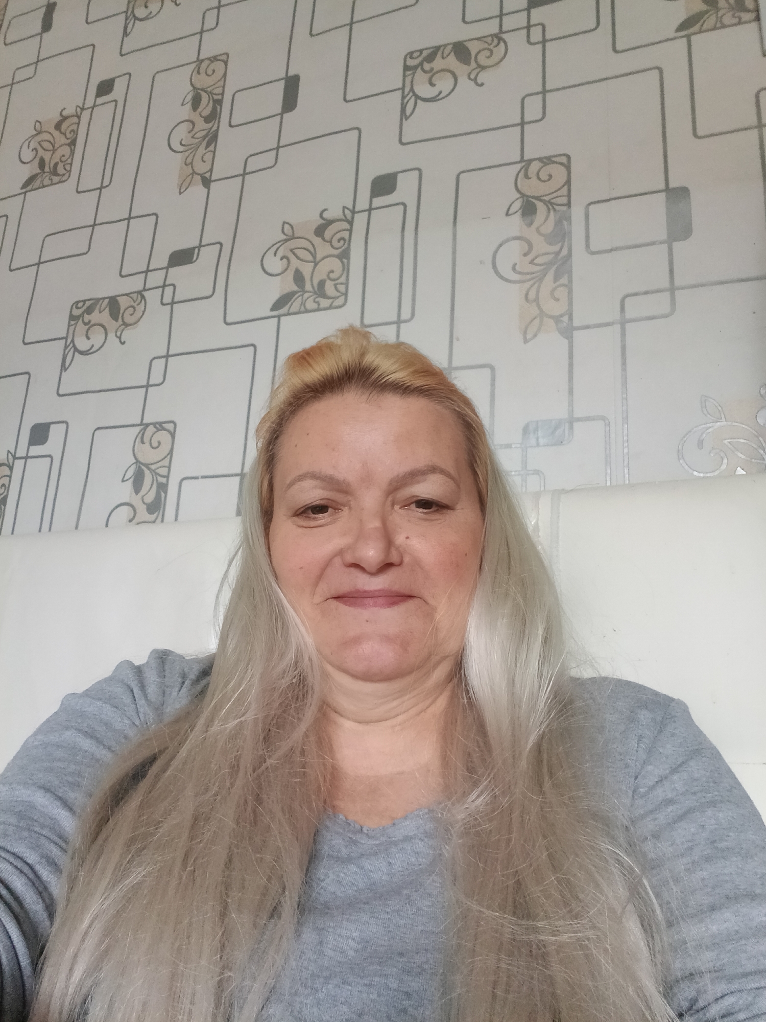 A woman is centered in the frame, seated and facing the camera. She has fair skin, blonde hair with darker roots, and a slightly smiling expression. She is wearing a gray, long-sleeved shirt. The background consists of a patterned wallpaper, featuring geometric shapes and floral designs, and a white headboard. The image appears to be taken indoors. There is no verifiable evidence to support that the location is in the city of Красный Маяк, Russia.