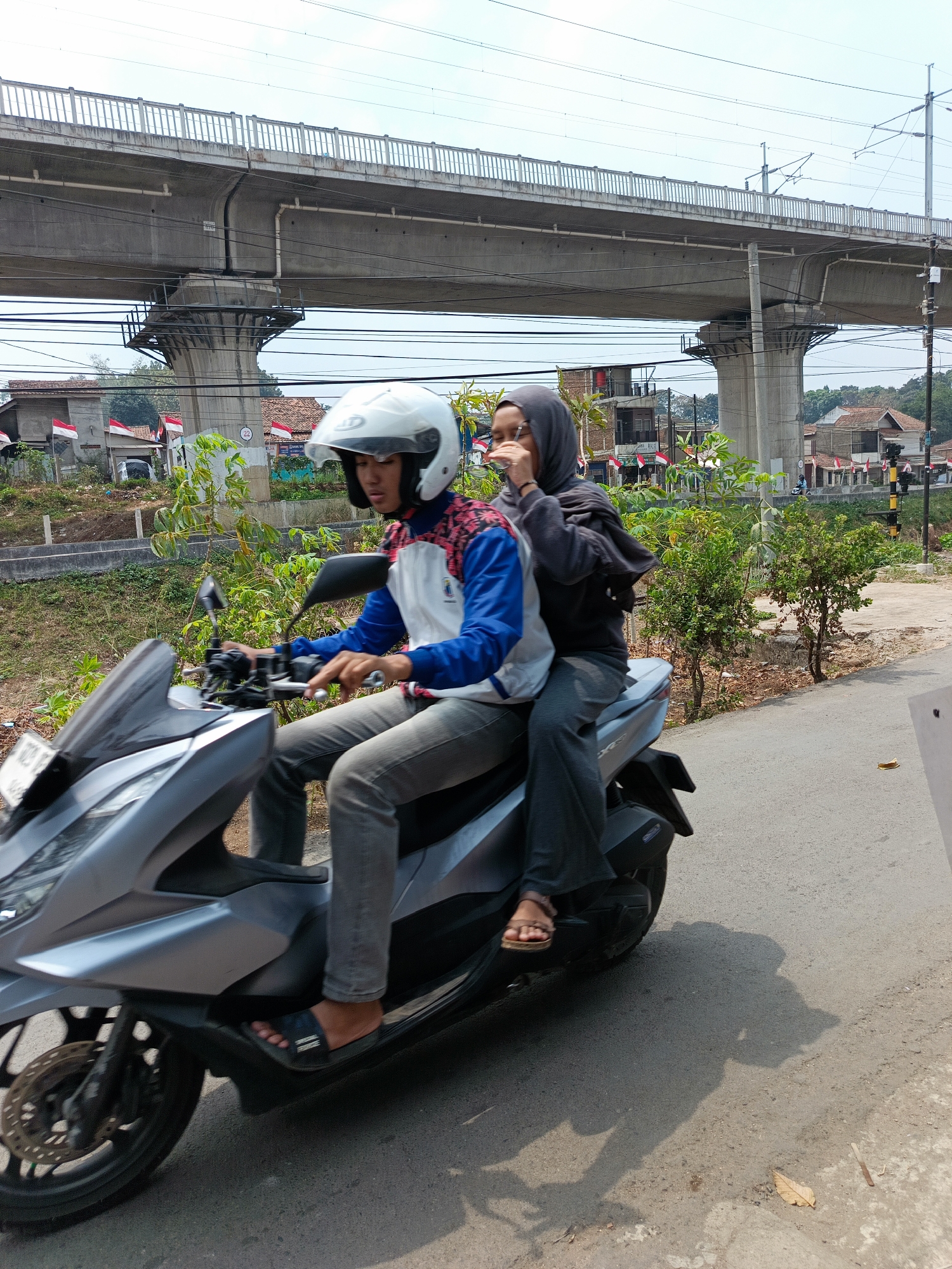 In Margajaya, Indonesia, two individuals are riding a silver scooter under a large concrete overpass. The person in front, wearing a white helmet and a blue and white jacket with red accents, is operating the scooter. The passenger behind, wearing a grey hijab and a dark grey jacket, is holding onto the driver and has their right hand near their face. The scooter is on a paved road. In the background, there are residential buildings with Indonesian flags displayed and lush green vegetation. The sky is clear and bright.