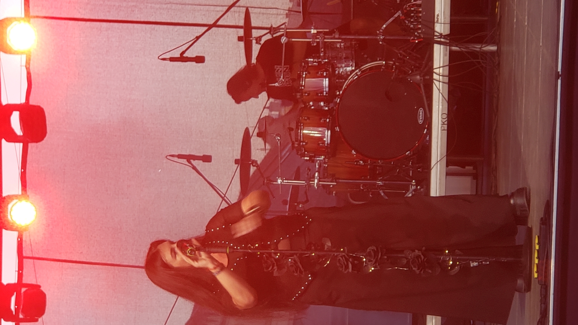 The image shows a live musical performance on an outdoor stage, likely in Prešov, Slovakia. A female vocalist, dressed in a black long skirt and a studded top adorned with black roses, stands center-left, singing into a microphone. Her long, dark hair cascades down her back. To the right of the vocalist, a drummer is seated behind a drum kit, playing. The stage is illuminated by bright red stage lights from above. Microphones are positioned on stands in front of the vocalist. A white banner with the text "PKO" is visible on the stage's front support structure. The overall scene is vibrant, with the red lighting casting a strong, atmospheric glow.