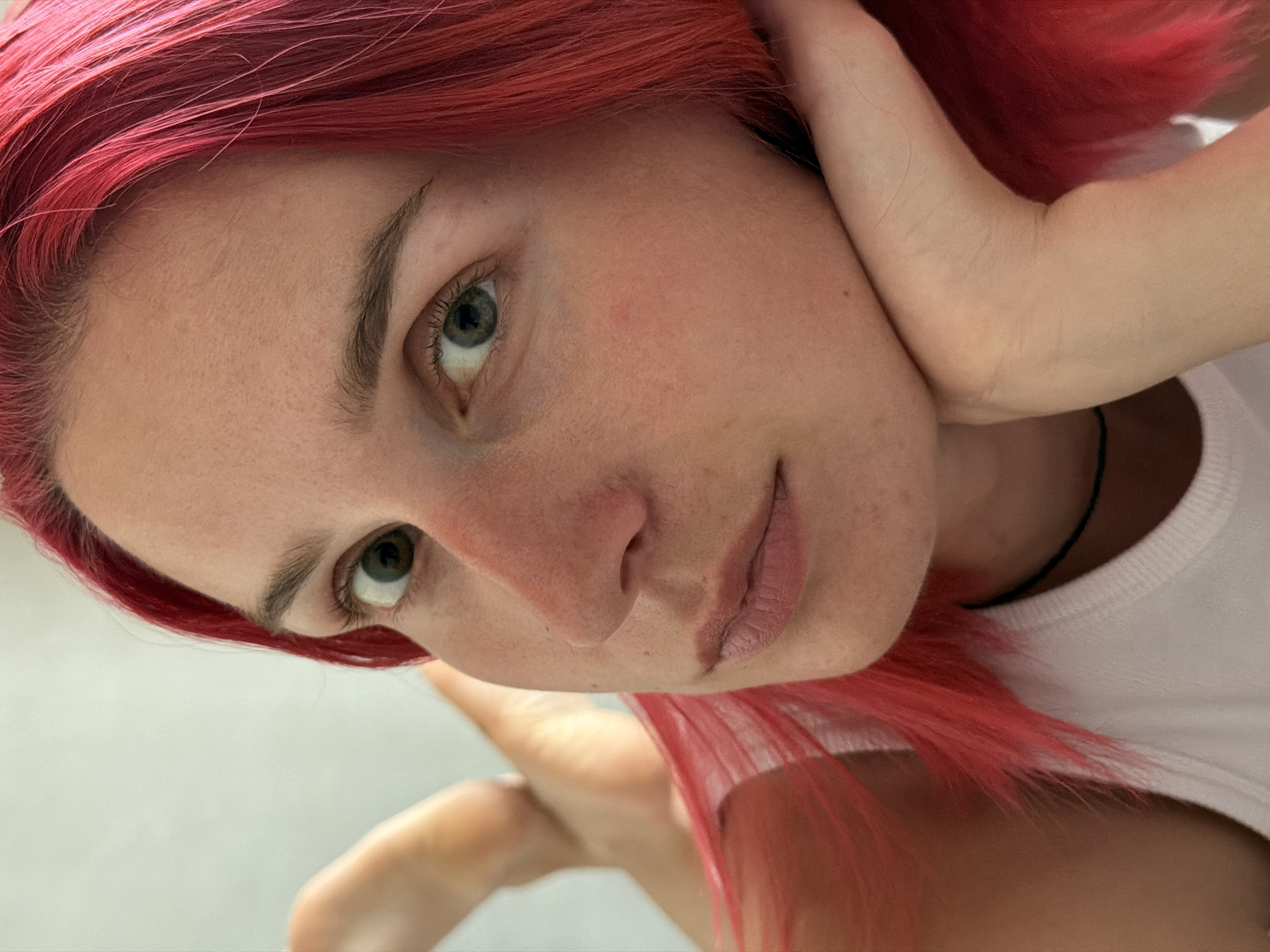 This is a close-up, portrait-style photograph of a young woman with vibrant, bright red hair, looking towards the camera. She is lying down, with her head resting on her hand. Her left arm is bent, and her hand supports her cheek, with her fingers gently splayed. Her right leg is bent at the knee and raised, with her foot visible in the upper left portion of the image. She is wearing a white tank top with a thin black cord or necklace visible around her neck. The background is a plain, light-colored surface, possibly a wall or a sheet. The lighting is soft, highlighting the texture of her skin and the strands of her hair. The scene is indoors, with no identifiable landmarks indicating a specific location within Chaikovsky, Russia.