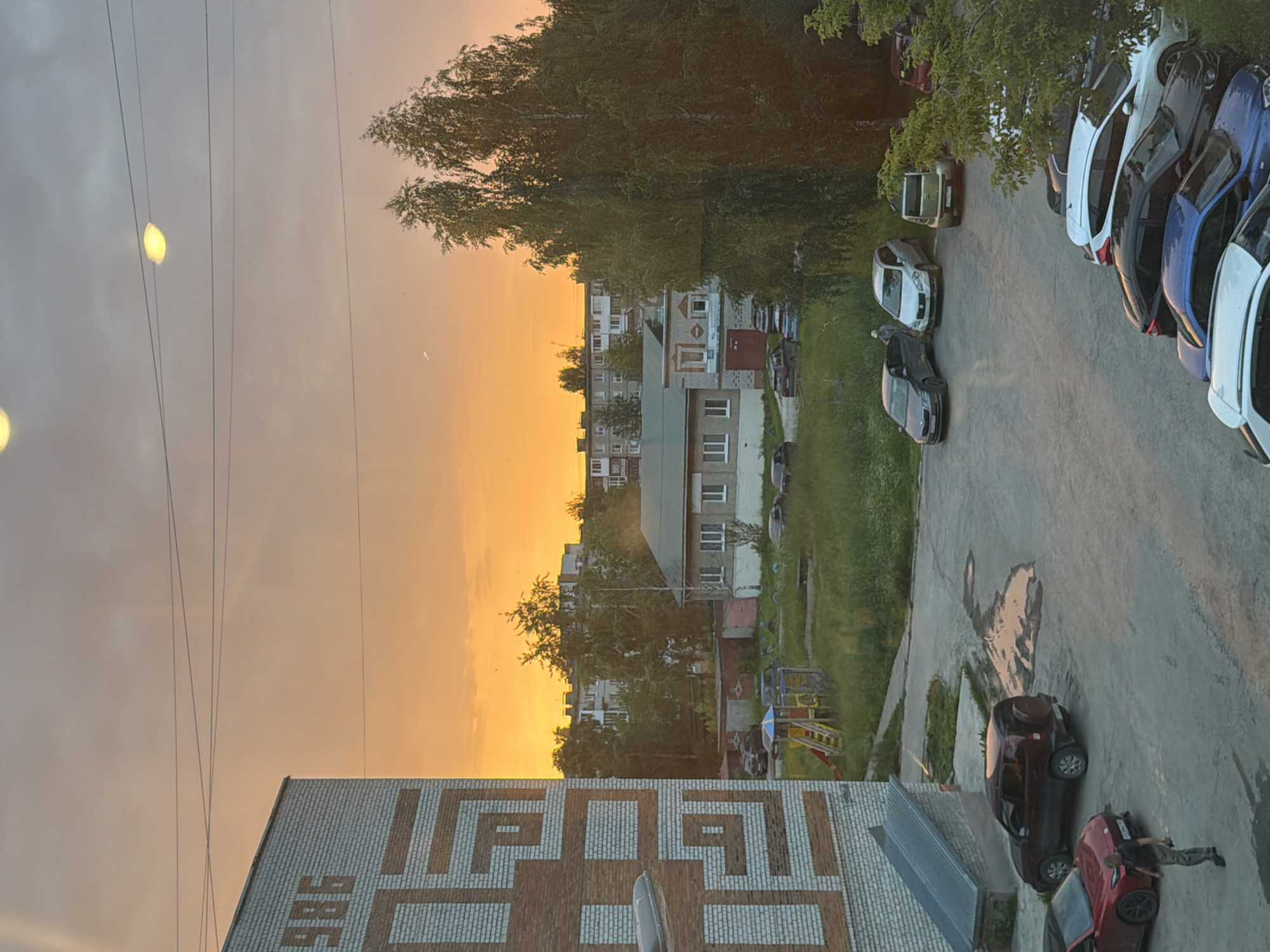 A wide shot captures a sunset over a residential area in Solikamsk, Russia. In the foreground, a brick building with the number "1986" on its side is visible on the left. A paved area with several parked cars, including a red sedan and a dark SUV, occupies the lower portion of the image. A person is seen walking away from the red car. Further back, a playground with colorful equipment is situated in a grassy area. Behind the playground, a low-rise building with a green roof and white-framed windows stands. A line of trees and taller apartment buildings form the backdrop, silhouetted against the vibrant orange and yellow hues of the sunset sky. Power lines are visible stretching across the upper portion of the frame.