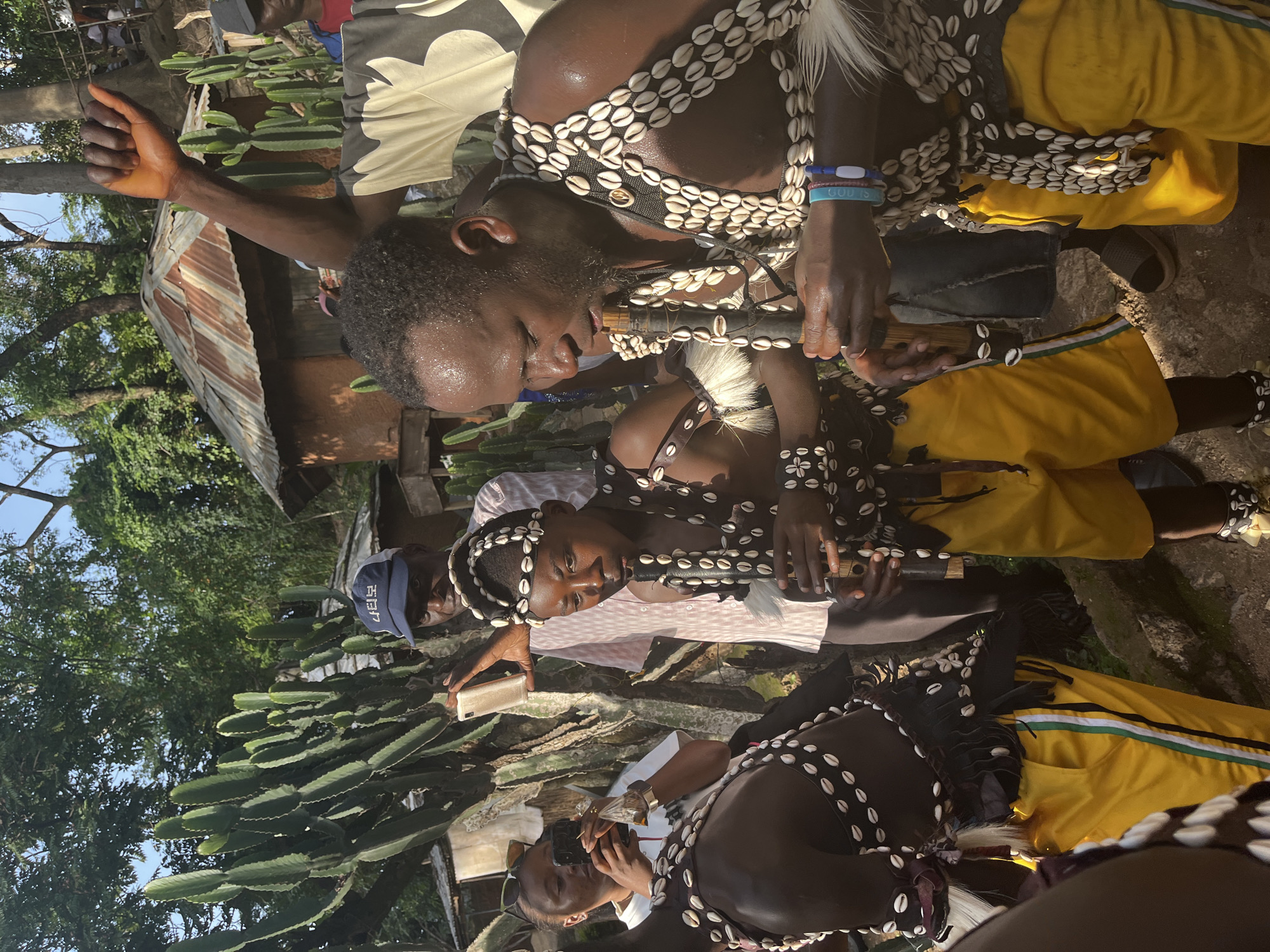 This is a medium shot of several people, some of whom appear to be participating in a cultural ceremony or performance. The subjects are primarily young Black individuals wearing traditional attire adorned with numerous cowrie shells. Some are wearing dark fabric vests and headdresses, also embellished with shells, and yellow skirts or trousers. One individual is playing a flute-like instrument.
In the background, there are lush green trees and a rustic-looking structure with a corrugated metal roof. Several large, green cactus-like plants are prominently visible in the foreground and midground. The lighting suggests it is daytime, possibly a sunny day given the shadows. The overall atmosphere appears to be lively and engaged, with some individuals looking directly at the camera or actively participating in the event. The presence of mobile phones being used by some individuals indicates a modern context within what seems to be a traditional setting.
There is some visible text on a blue baseball cap worn by one of the individuals. The text appears to be in Korean, with characters that read "나비야". One of the individuals also has bracelets on their wrist with text that appears to say "GOD IS LOVE".
The scene is set in Jos, Nigeria, as indicated by the location context provided.