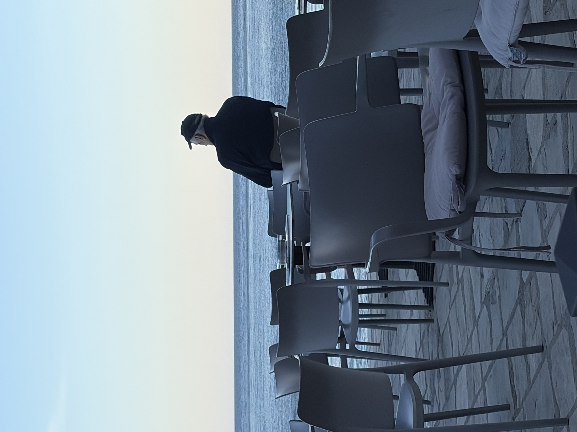 The image depicts an outdoor seating area, likely a café or restaurant, overlooking a vast body of water, possibly the sea, consistent with a coastal location like Budva, Montenegro. The setting appears tranquil, with a clear sky transitioning from a soft blue overhead to a pale yellow-orange near the horizon, suggesting either early morning or late afternoon light.
In the foreground and midground, numerous contemporary-styled chairs, made of a grey or dark grey material, are neatly arranged in rows, all facing towards the water. Many of these chairs have light-colored, possibly white or cream, cushions on their seats. The ground beneath the chairs is paved with light-toned tiles or stone, exhibiting a geometric or interlocking pattern.
A single person, an older man, is visible standing near the edge of the seating area, with his back partially turned to the viewer. He is wearing a dark, long-sleeved top and a dark baseball cap. His gaze is directed outwards towards the calm, rippling water. The empty chairs imply a quiet moment, perhaps before opening hours or during an off-peak time, contributing to the serene atmosphere. No specific activity other than observation is discernible. The overall mood is one of peace and quiet contemplation, with the man seemingly lost in thought while admiring the scenic view. There is no visible text in the image.