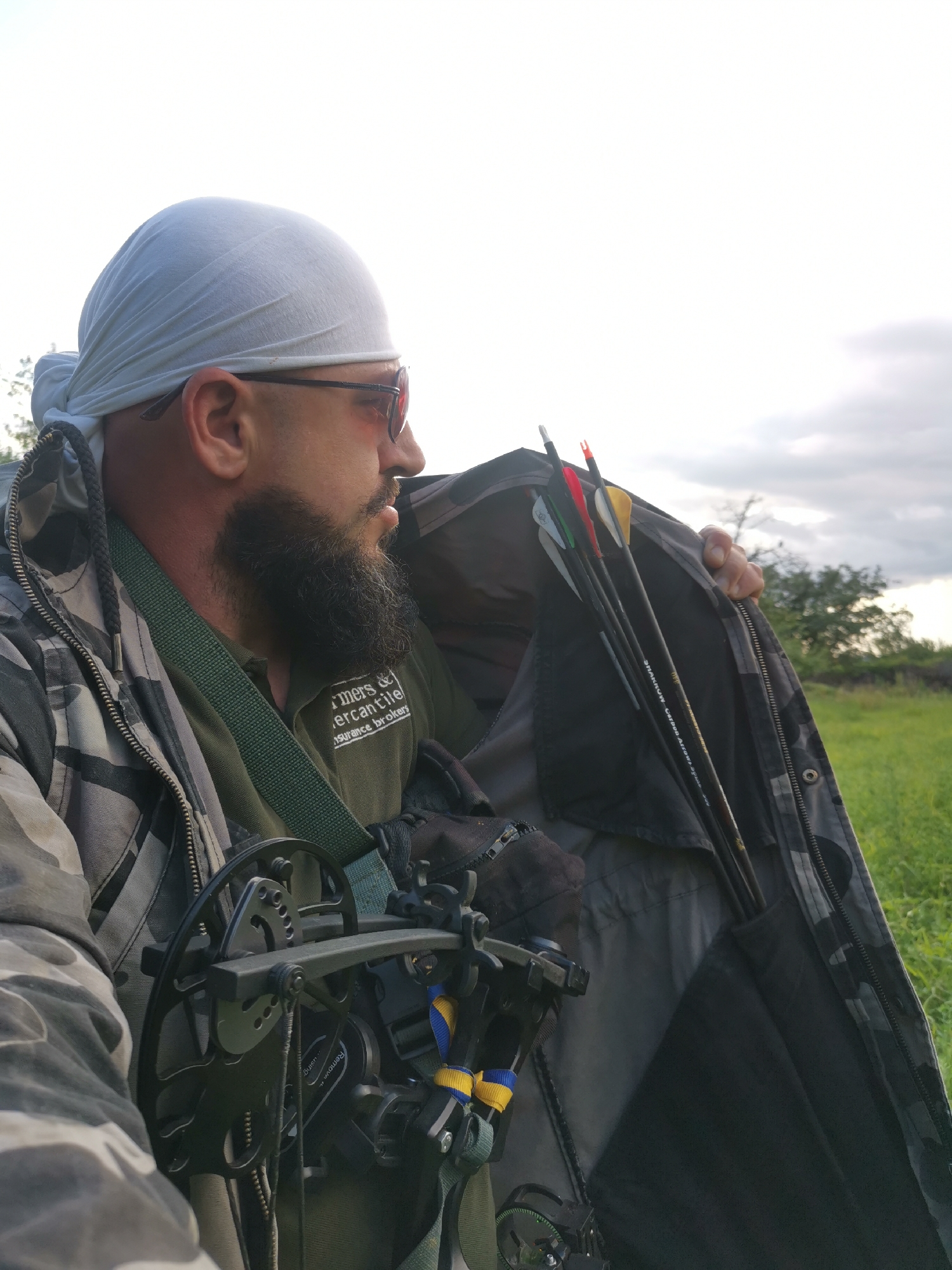 The media file features a man outdoors, with a dense beard and a light-colored bandana covering his head. He is wearing sunglasses and a camouflage jacket over a green shirt. The shirt has visible text which appears to be a logo: "Farmers & Mercantile Insurance Brokers". The man is holding a compound bow, attached to his chest by a strap. Inside the open jacket, several arrows are stored, some with colored fletching. The scene appears to be a grassy field with some trees in the background, suggesting a rural or natural setting. The lighting indicates it is daytime, possibly late afternoon or early evening, with overcast or cloudy skies. The man's gaze is directed away from the camera, towards the right side of the frame, giving him a thoughtful or focused expression. The overall impression is of someone engaged in outdoor activities, possibly hunting or practicing archery. The location context is Semypolky, Ukraine.