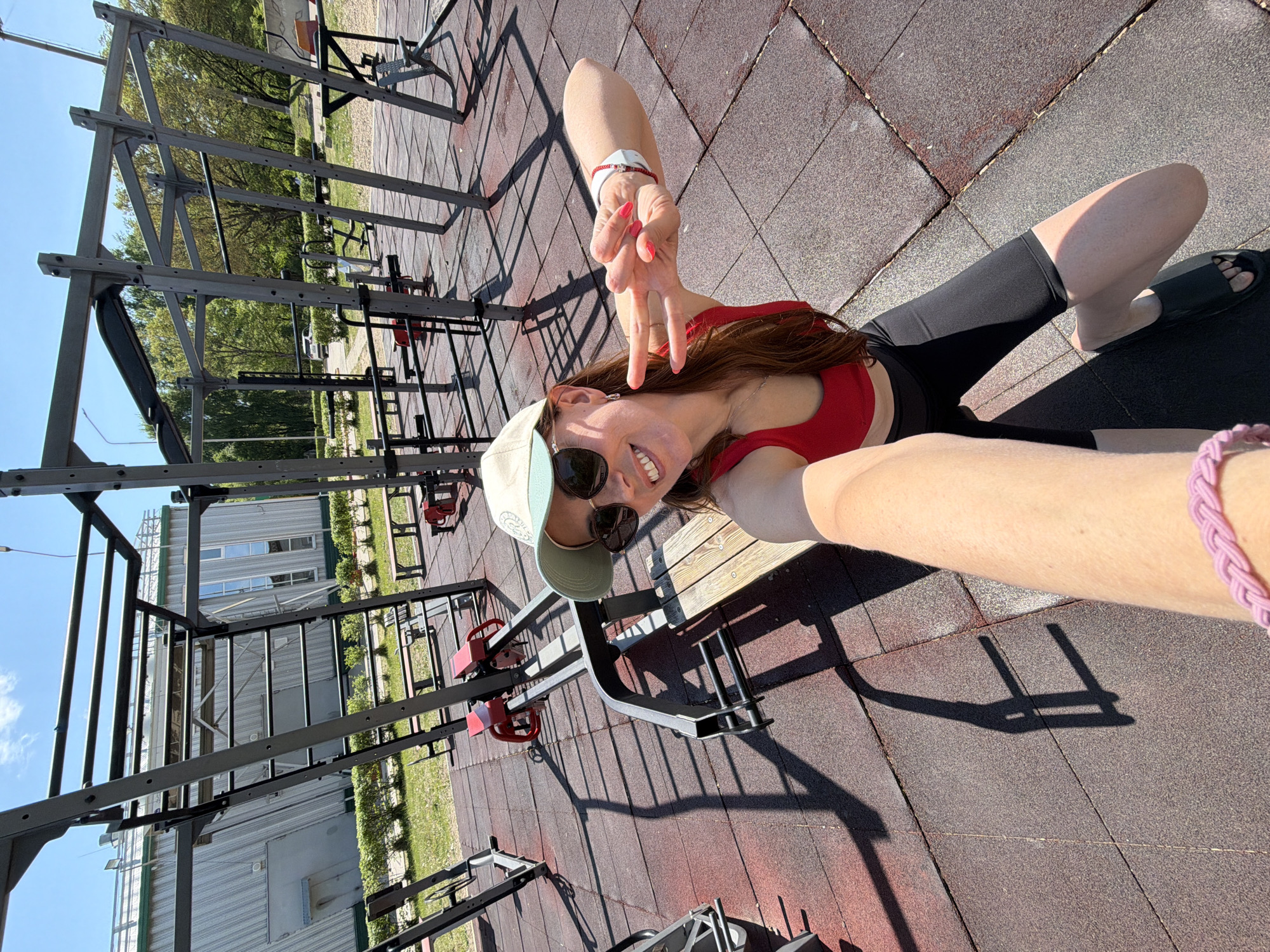 A woman is lying on a red and grey tiled surface at an outdoor fitness park in Birobidzhan, Russia. She is wearing a red crop top, black shorts, a light green baseball cap, and sunglasses. She is holding her hand up in a peace sign. In the background is a metal jungle gym, a building with windows, and green trees. The sky is blue and sunny.