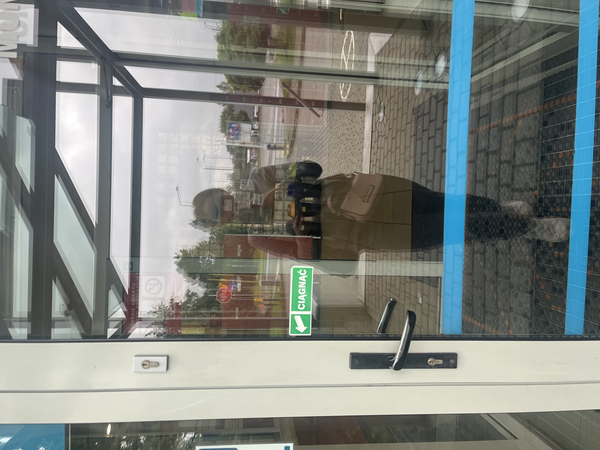 The image shows the interior of a glass-enclosed entryway with a person reflected in the glass. The person, wearing a mask and a light-colored jacket, is holding a pink purse. Outside the entryway, a street with traffic lights and trees is visible. A green sign with a white arrow and the word "CIĄGNĄĆ" (Polish for "pull") is attached to the door. A black door handle and a keyhole are also visible on the door. The floor inside the entryway is a dark mat with blue tape across it, and the floor outside is paved with bricks.