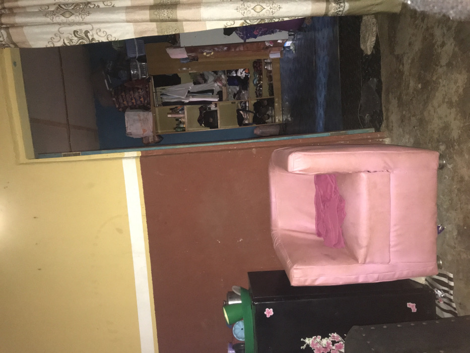 An interior view in Oba-Ile, Nigeria, depicts two adjoining rooms. The foreground room features a bi-color wall, with an upper yellow section and a lower reddish-brown section, separated by a white horizontal stripe. The concrete floor is soiled. A black compact refrigerator or cabinet stands on the left, adorned with pink floral stickers. On its top rests a green plastic tray containing various containers, including a blue plastic sieve. To the right of the refrigerator is a pink single armchair, over which a folded red or pink fabric is draped. The armchair is supported by four metallic legs. Beneath the refrigerator, a black and white patterned mat is visible.
An open doorway on the right leads to a second room. A striped curtain, beige and white, partially obscures the doorway on the right. The second room has blue walls and a darker floor, possibly tiled. A wooden shelving unit or wardrobe dominates the left side of this room, containing numerous items including bottles, bags, and shoes on shelves, and various garments hanging from a rail. Additional bags and containers are stacked on top of the shelving unit. The ceiling of this inner room reveals exposed wooden beams. No individuals are present in either room, and no actions or interactions are occurring.