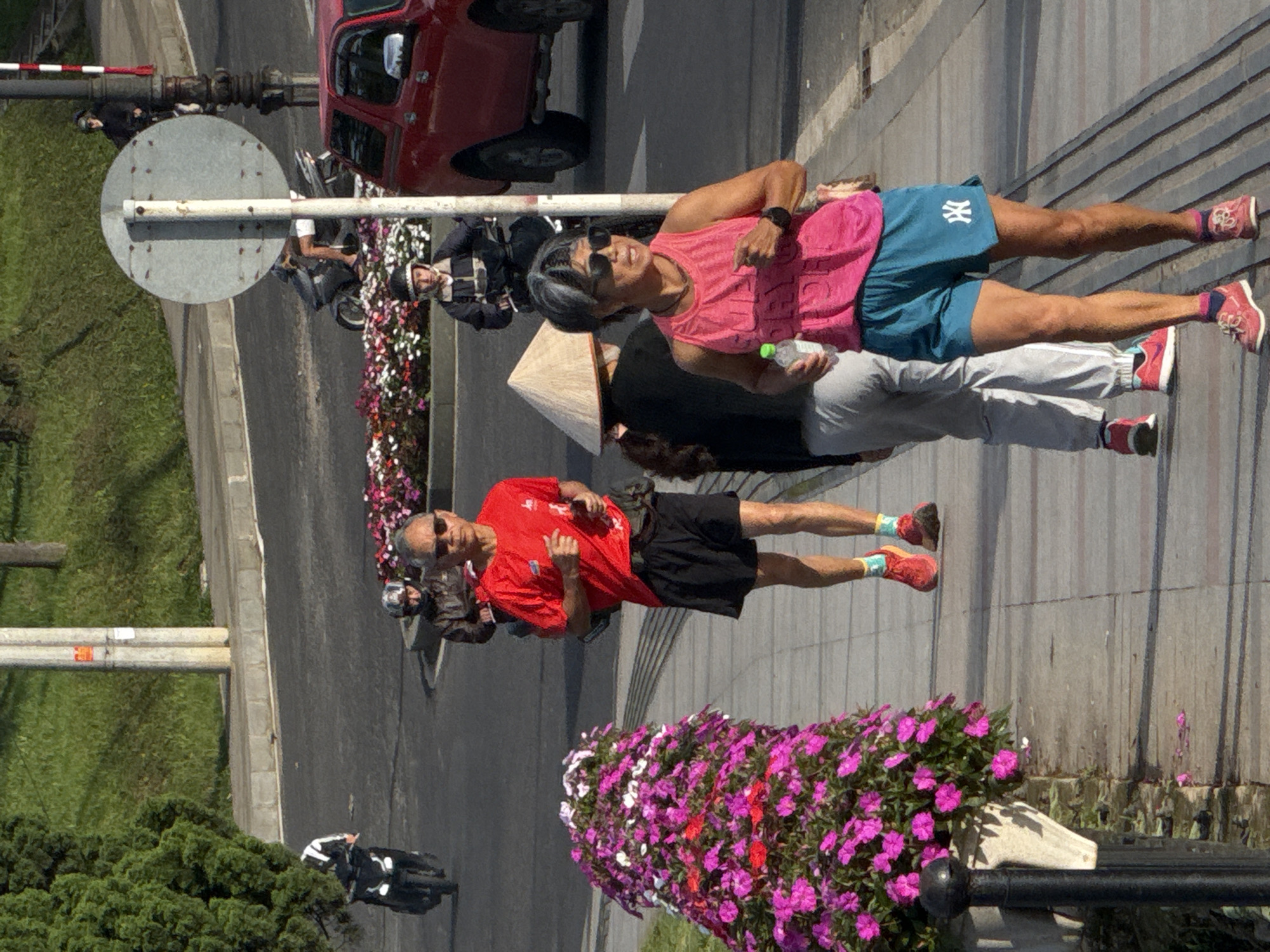 The image depicts an outdoor urban scene with a paved walkway and a street, likely in Lâm Viên - Đà Lạt, Vietnam, given the presence of flowering plants and architecture. A man in a red t-shirt and black shorts is jogging on the walkway. To his right, a woman in a pink tank top and teal shorts is also jogging, holding a water bottle. Behind them, another individual wearing a conical hat and a dark top is walking. In the background, several people are on motorcycles on the street, and a red truck is parked. The walkway is bordered by a vibrant display of pink and white flowers. The overall impression is a sunny day with people engaged in outdoor activities.